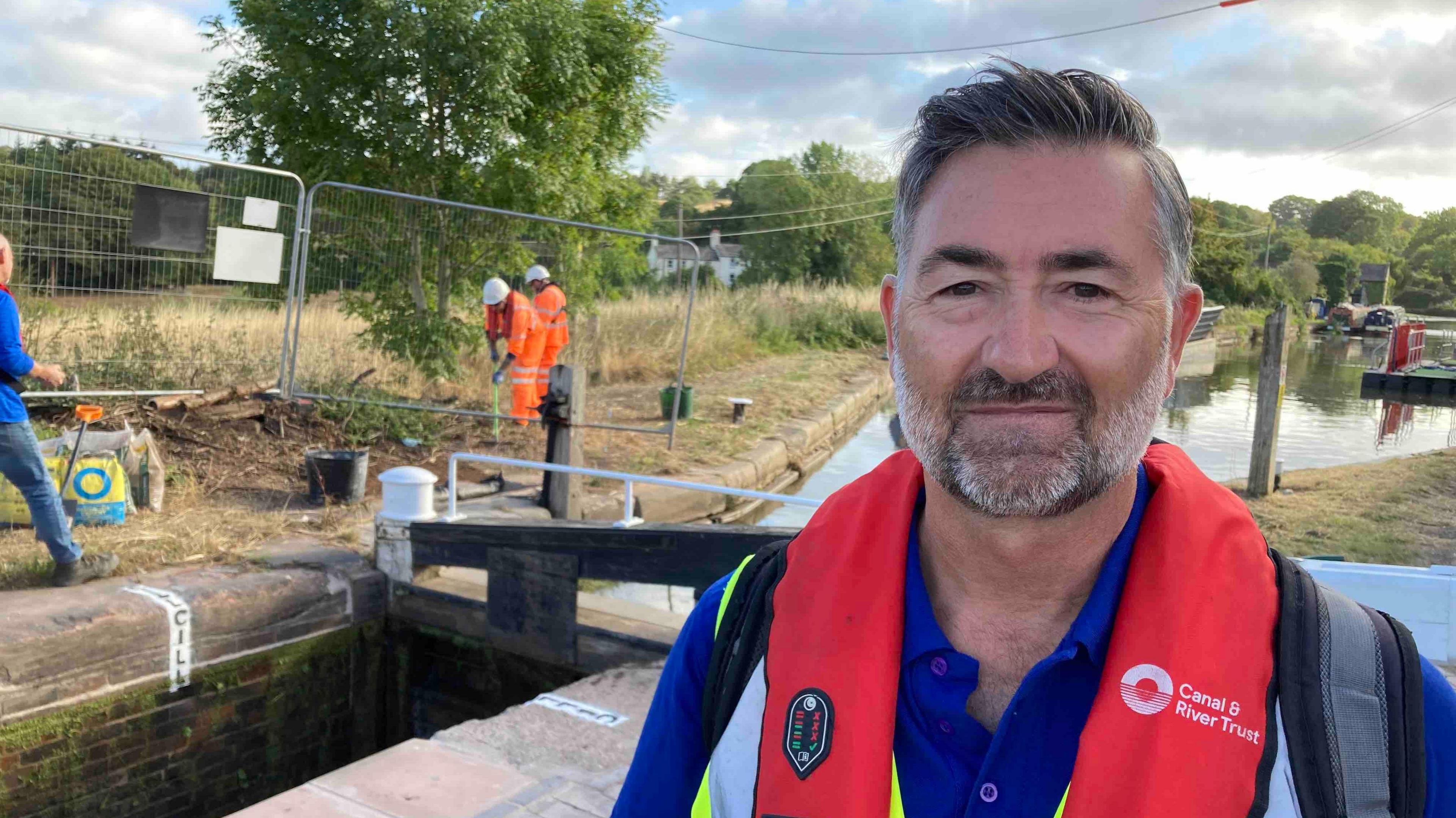 Neil Bedford wearing a red life jacket is stood next to the locks with other workers in the background. He has short dark greyish hair and a grey beard.