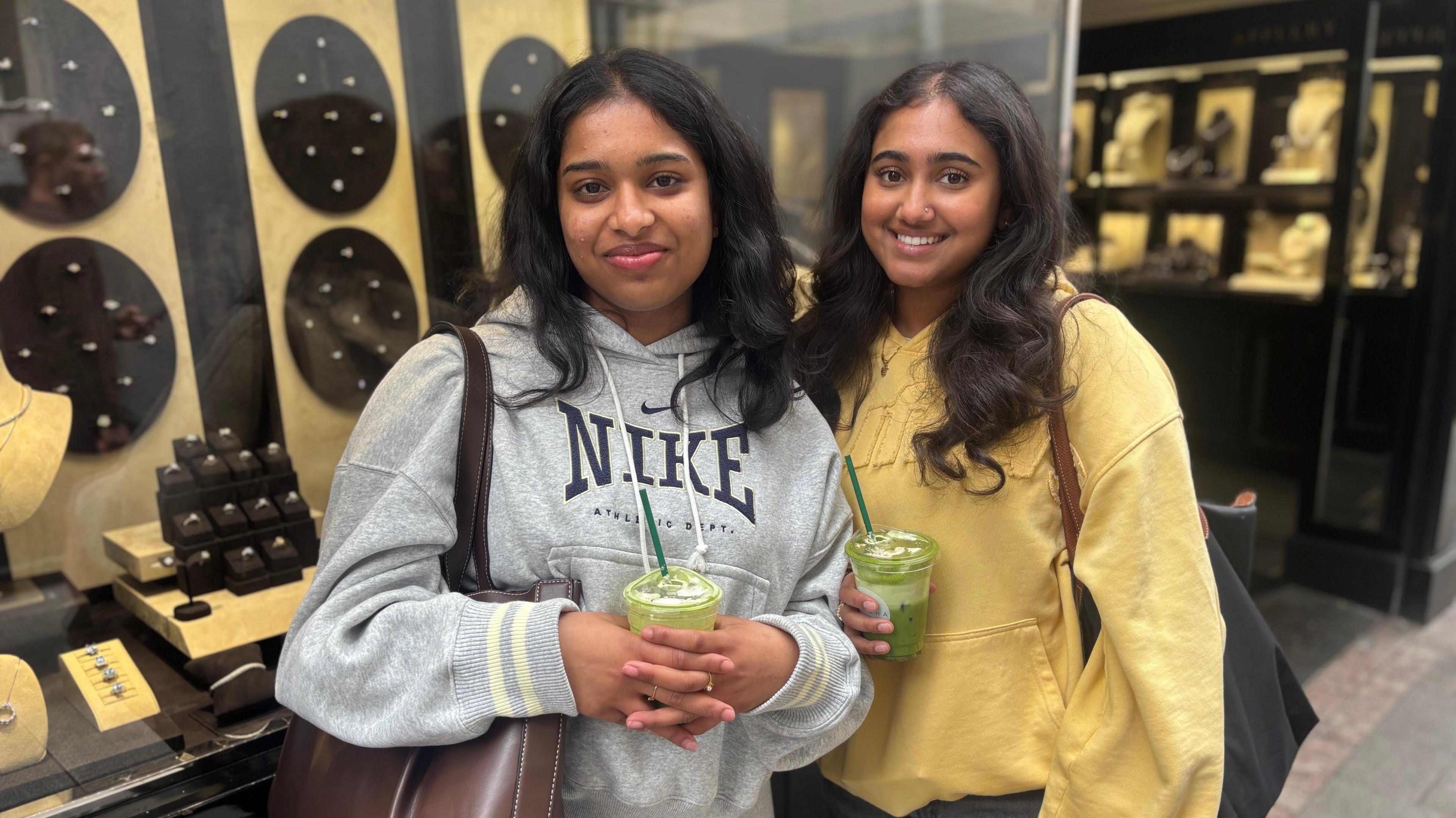 Two girls wearing grey and yellow hoodies stand in front of a jewellery shop 
