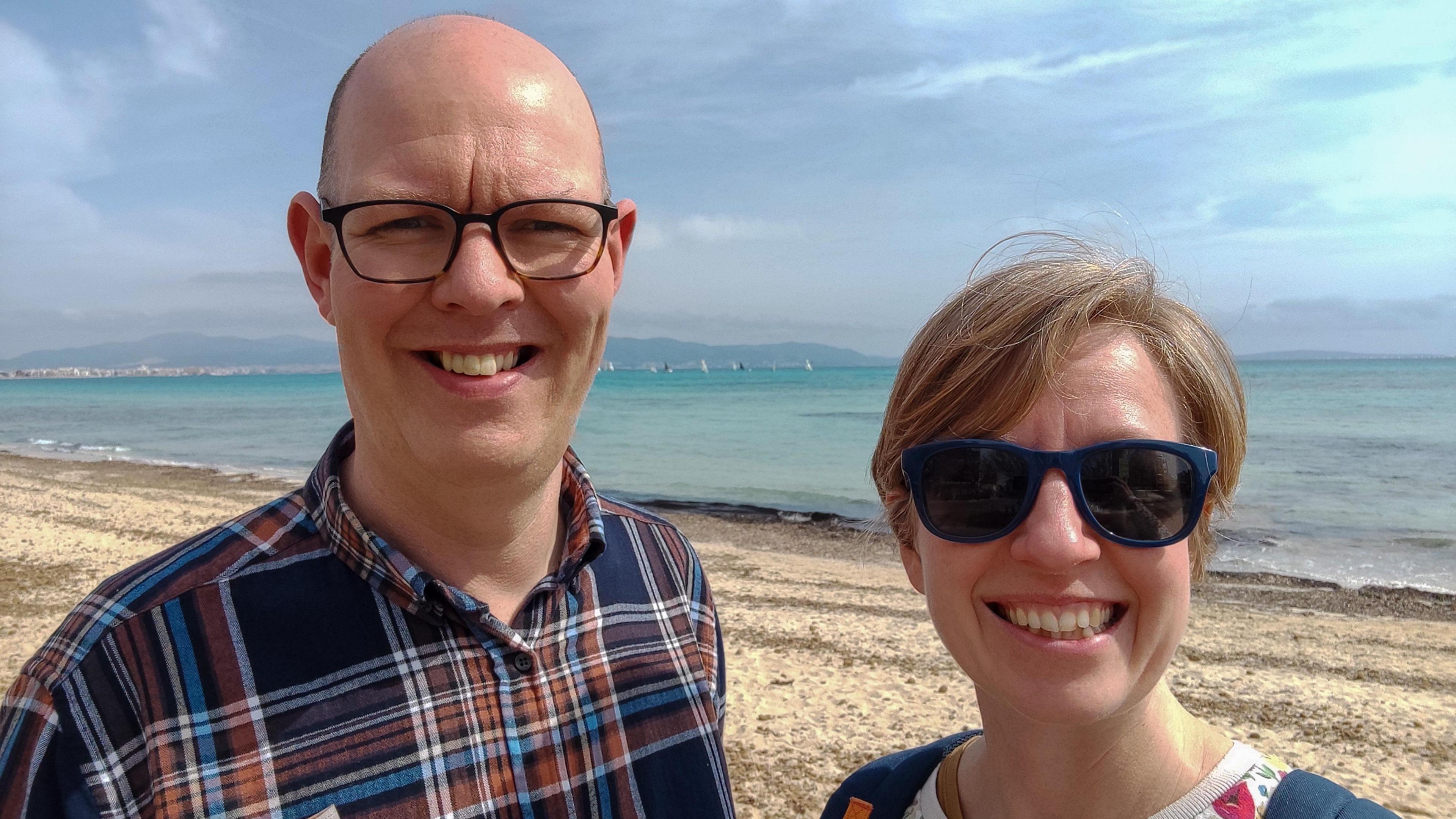 A man with a shaved head and wearing dark framed classes and a blue and brown plaid shirt stands next to a woman with short blonde hair and sunglasses. They are stood on a sandy beach and behind them is the sea, with sail boats and a coastline in the distance.