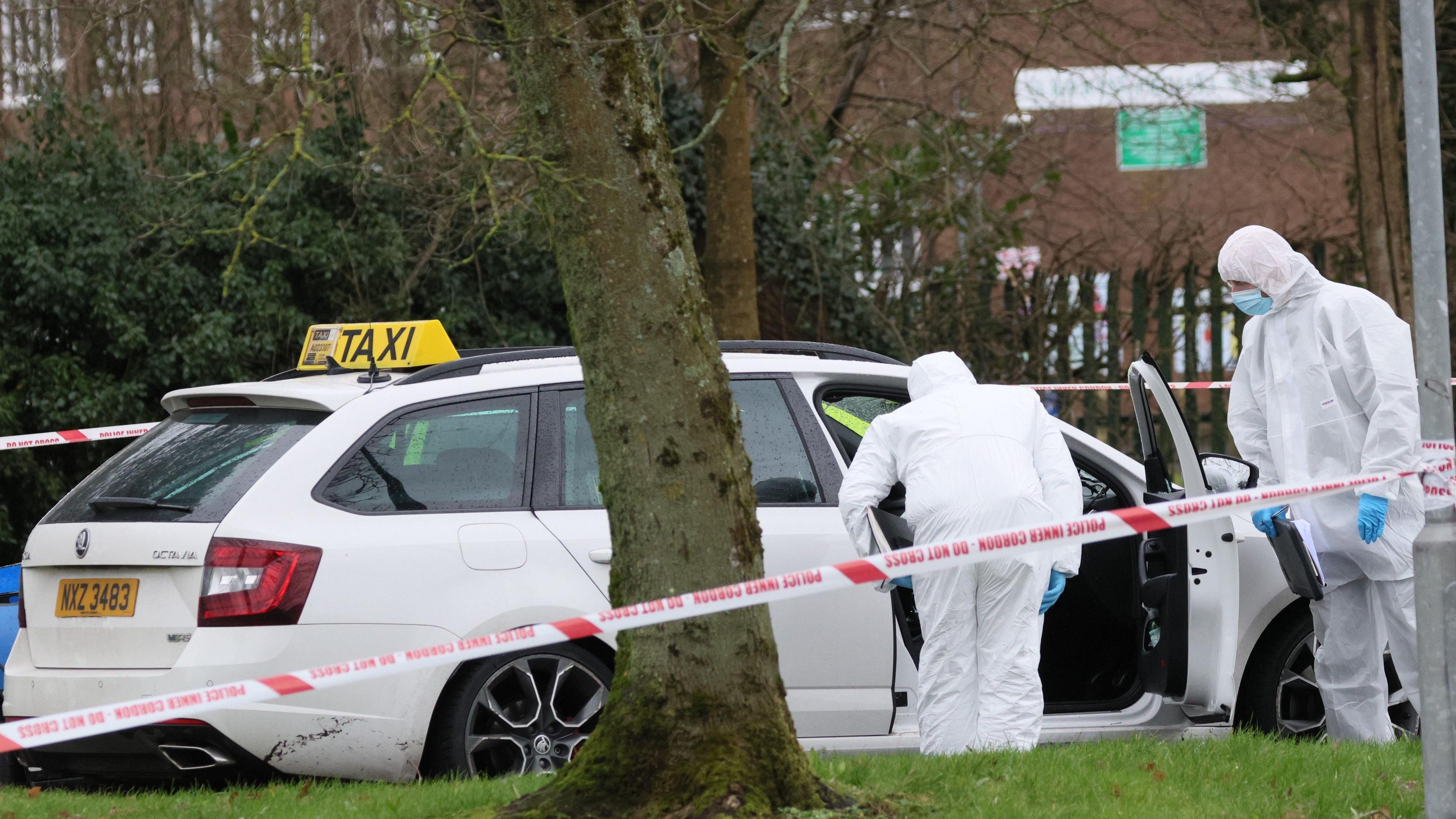 A police cordon with two officers in white forensic suits with blue gloves and face masks, thay are looking inside a white Skoda vehicle that has a yellow taxi sign above it. The word TAXI is in black.