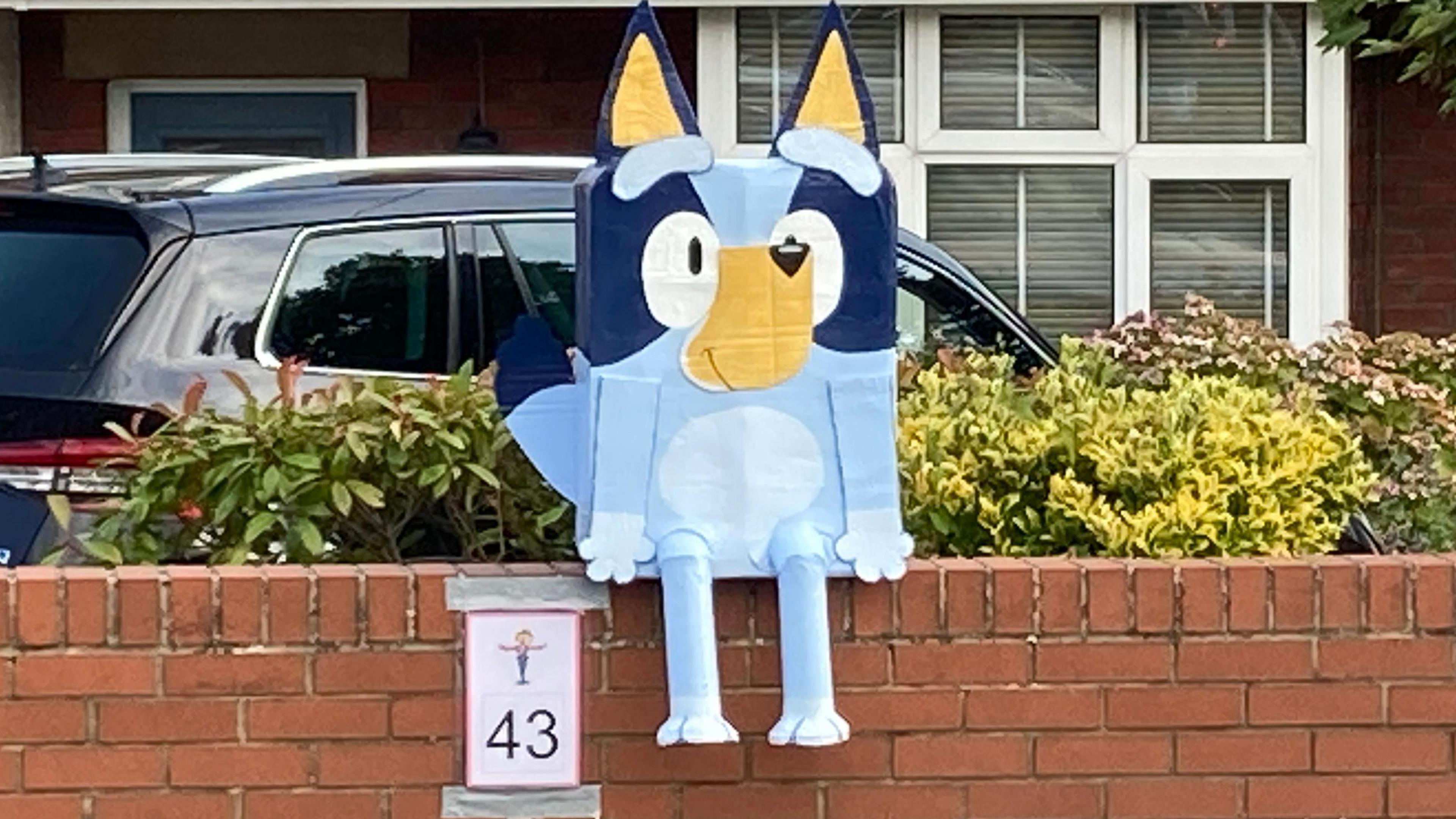 A carboard version of character Bluey sits on a front garden wall with his trail number