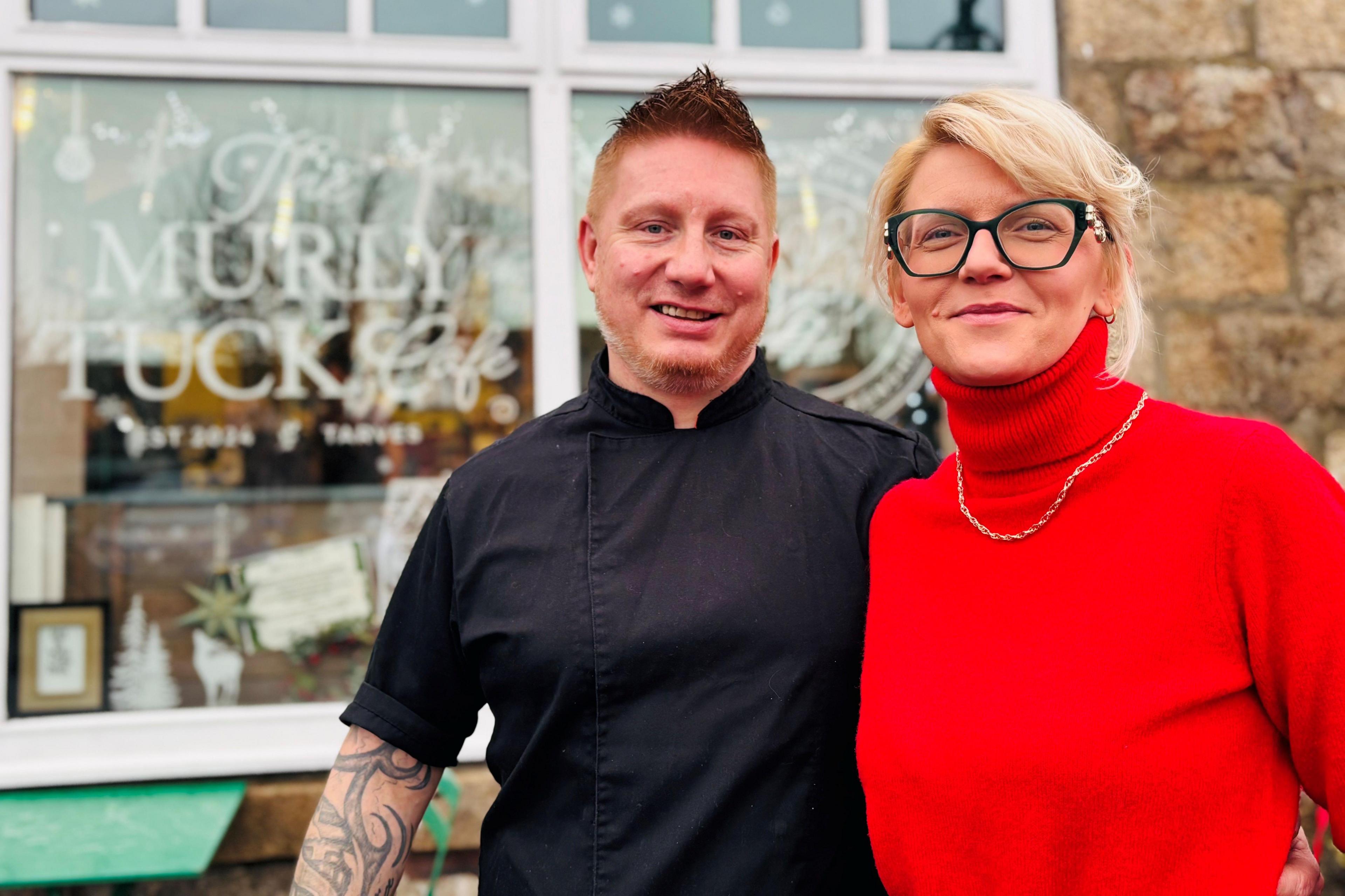 Photo of Lukasz and Edyta Kranz outside their cafe Murly Tuck in Tarves, he is wearing a black chef's top and she is wearing a red top.