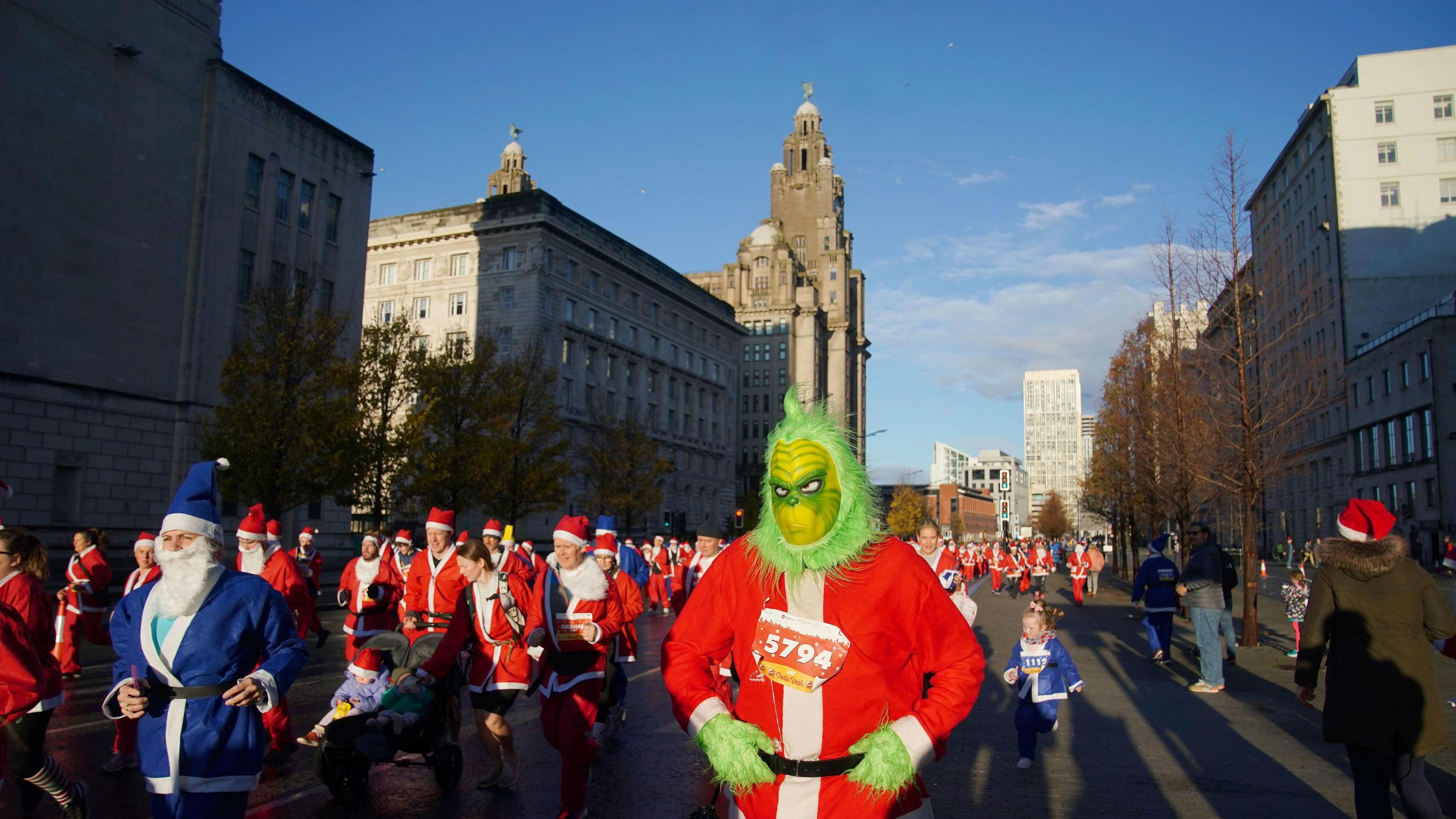 Masses of runners in mainly red Santa suits and some in blue Santa suits, including one wearing a green Grinch outfit with a Santa jacket over the top