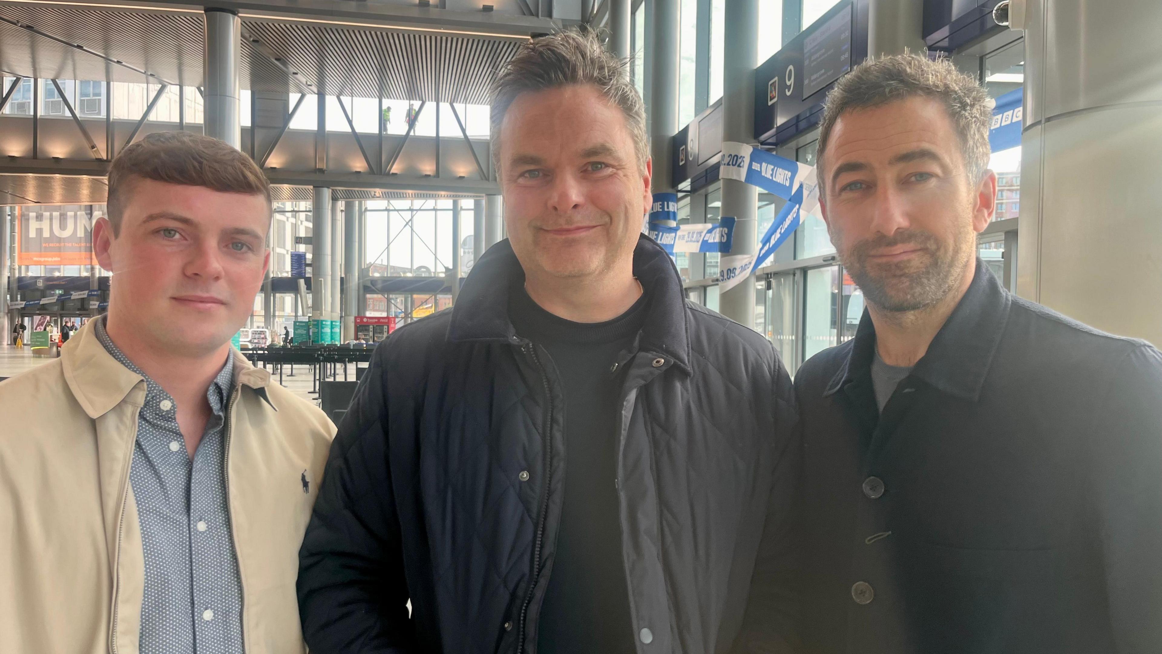 Nathan Braniff, Declan Lawn and Adam Patterson are stood in Belfast's Grand Central Station and are looking directly at the camera. Braniff on the left is a younger man with a cream jacket and a blue collared, buttoned shirt. Lawn in the centre is wearing a black padded coat and black shirt, and Patterson is wearing a dark buttoned coat.