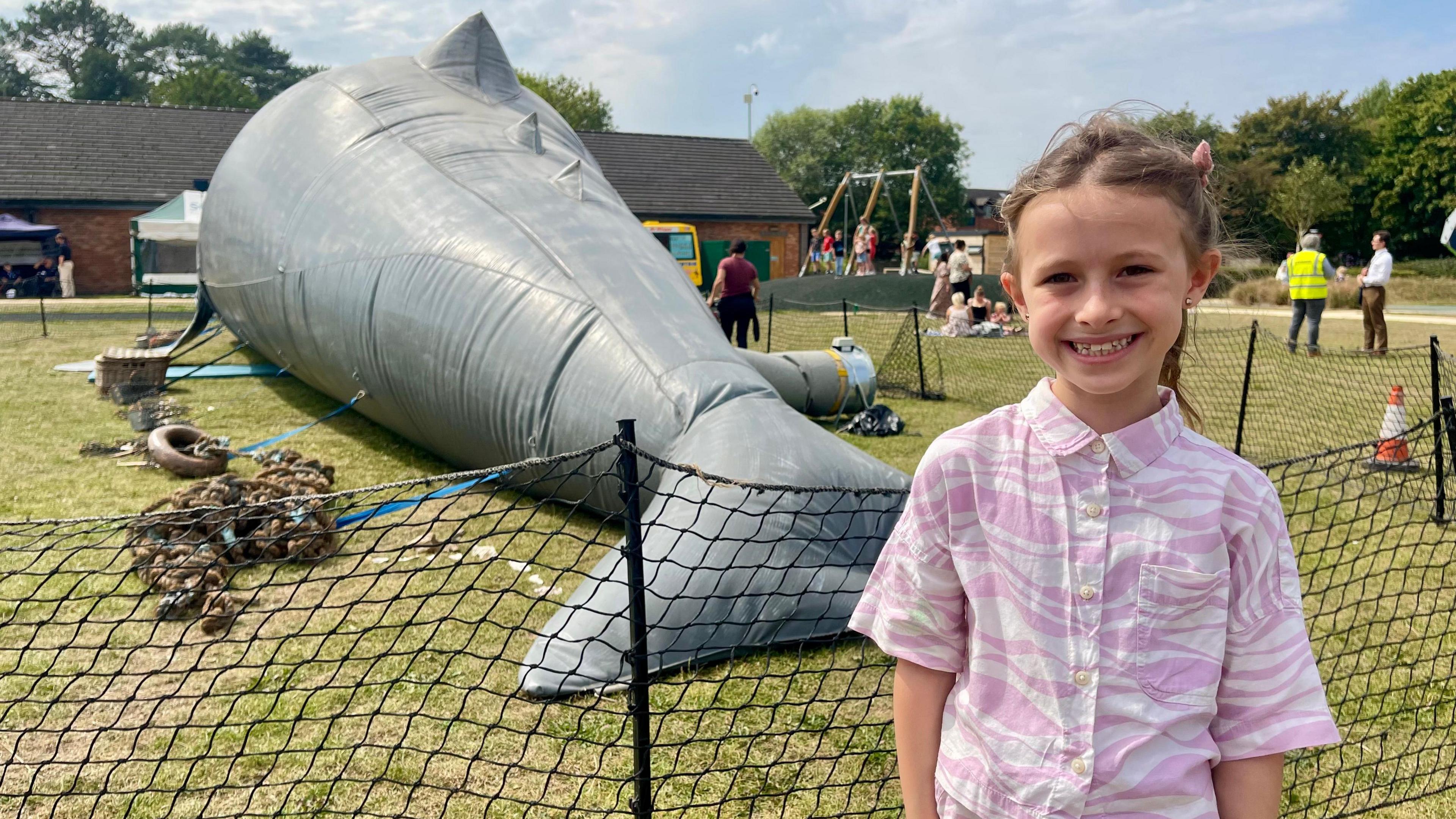 A girl with a purple and white striped shirt stands in front of an 18 metre long inflatable sperm whale in a park. Her brown hair is tied back and she is smiling. The whale is tied down by blue straps and has seaswept rubbish beside it. The whale is surrounded by black netting acting as a barrier. A brick building, gazebos, an ice cream van, swings and a traffic cone can be sen in the background.