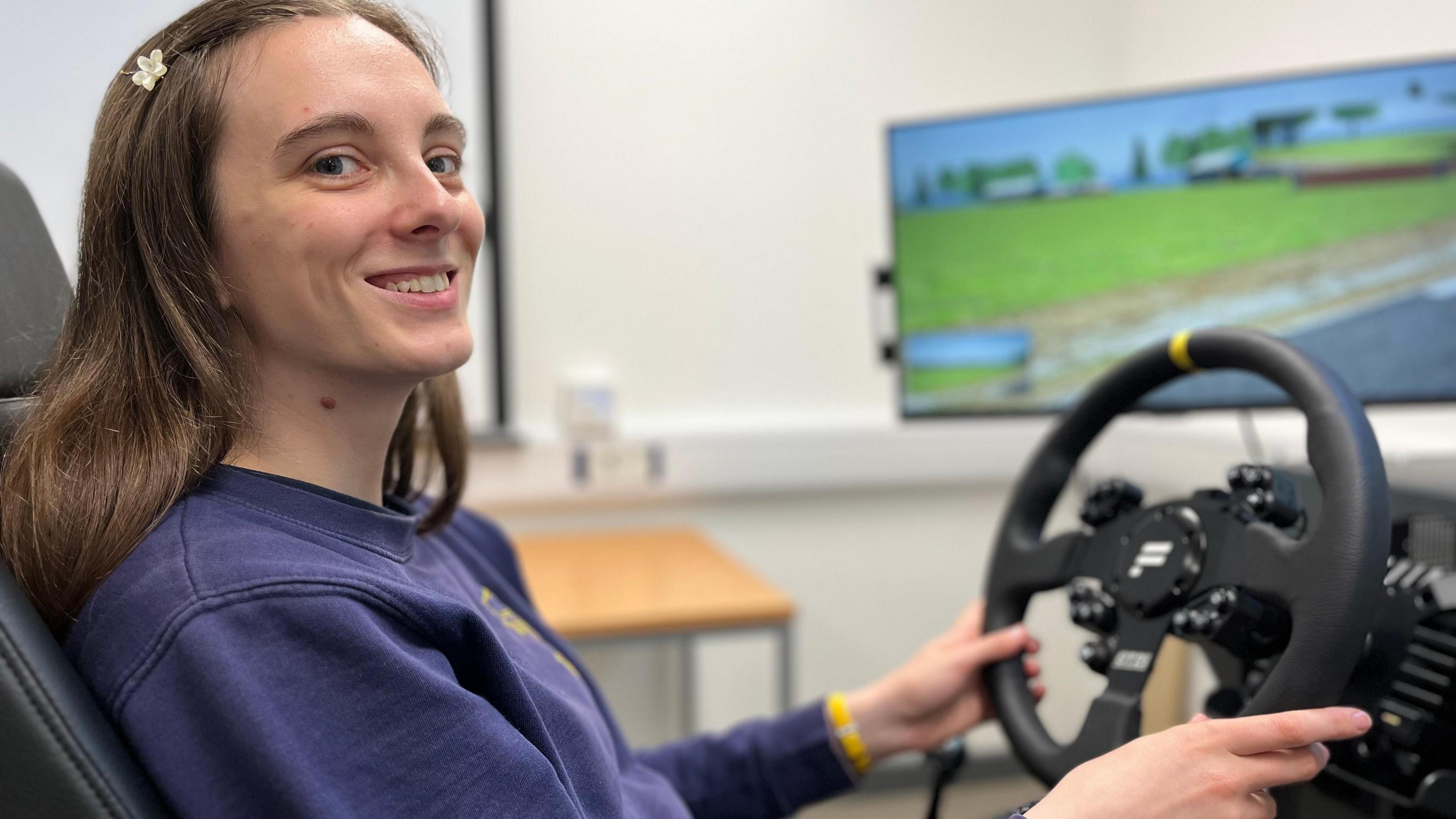 A girl with a navy blue jumper is sat in a gaming style chair in front of a large screen which show a driving simulation game. She is using a mock stirring wheel to drive. She has both hands on the stirring wheel and she is looking directly at the camera. She has dark brown hair with a small flower hair clip in her hair. She is also wearing a yellow watch on her left wrist.
