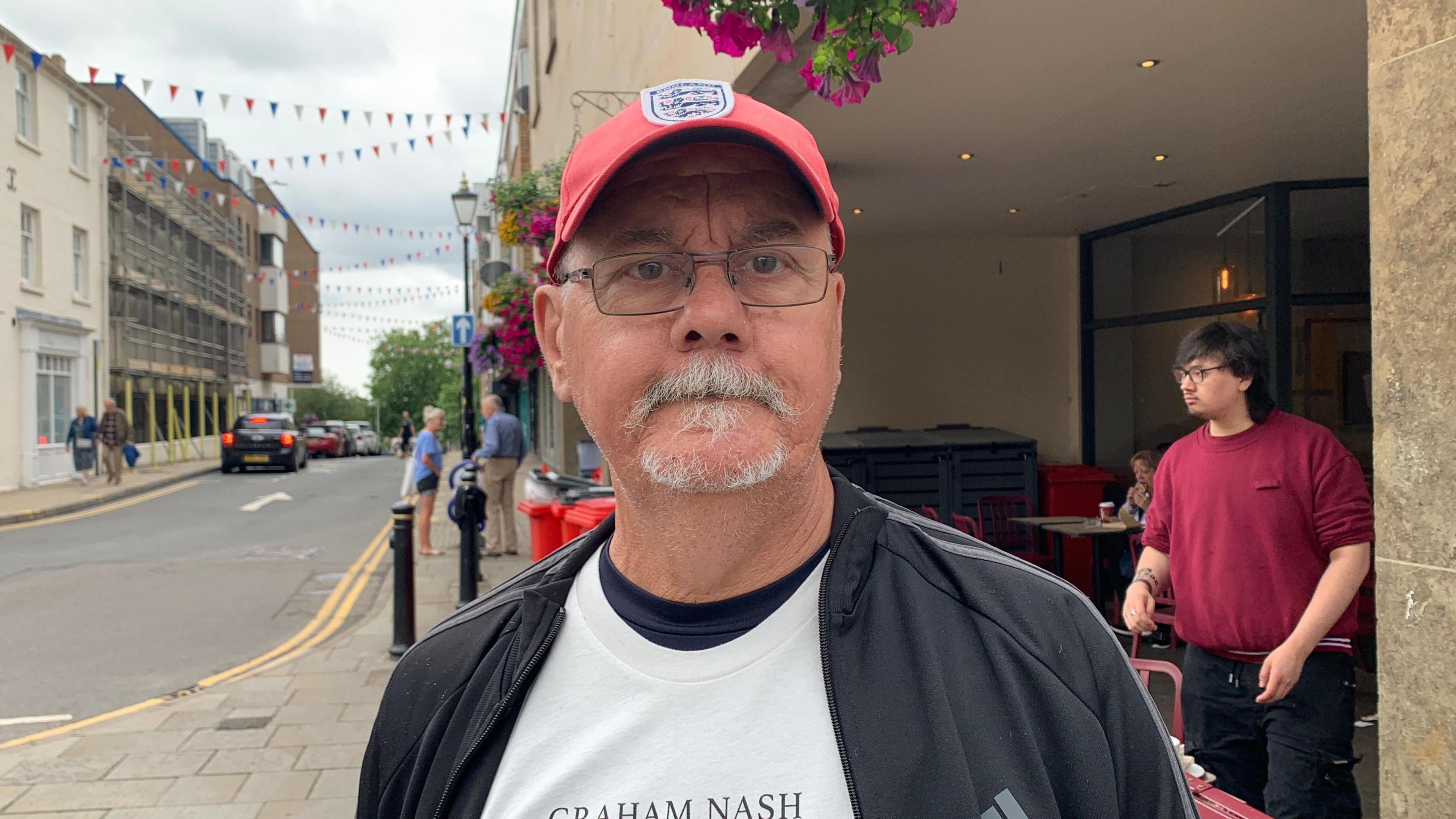 Geoffrey Manion looks at the camera. He has a great goatee beard. He is wearing clear glasses and a red baseball hat with the England football team's logo on it.