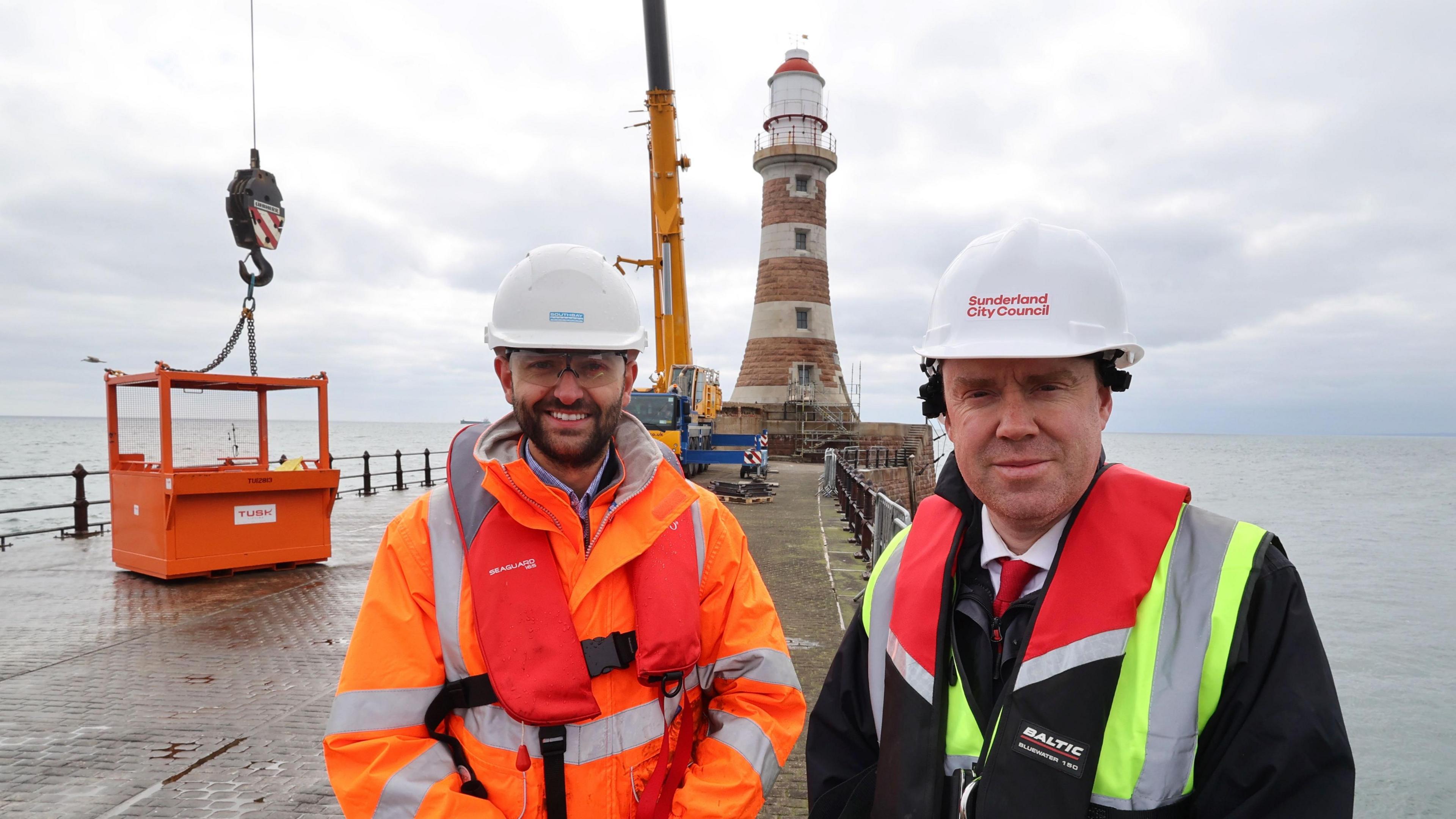 Benjamin Leng (left) and Councillor Kevin Johnston standing in front of the lighthouse on Roker Pier. Mr Leng is wearing an orange hi-vis outfit and has a dark beard. Mr Johnston is wearing a black jacket. Both men are also wearing white hard hats.