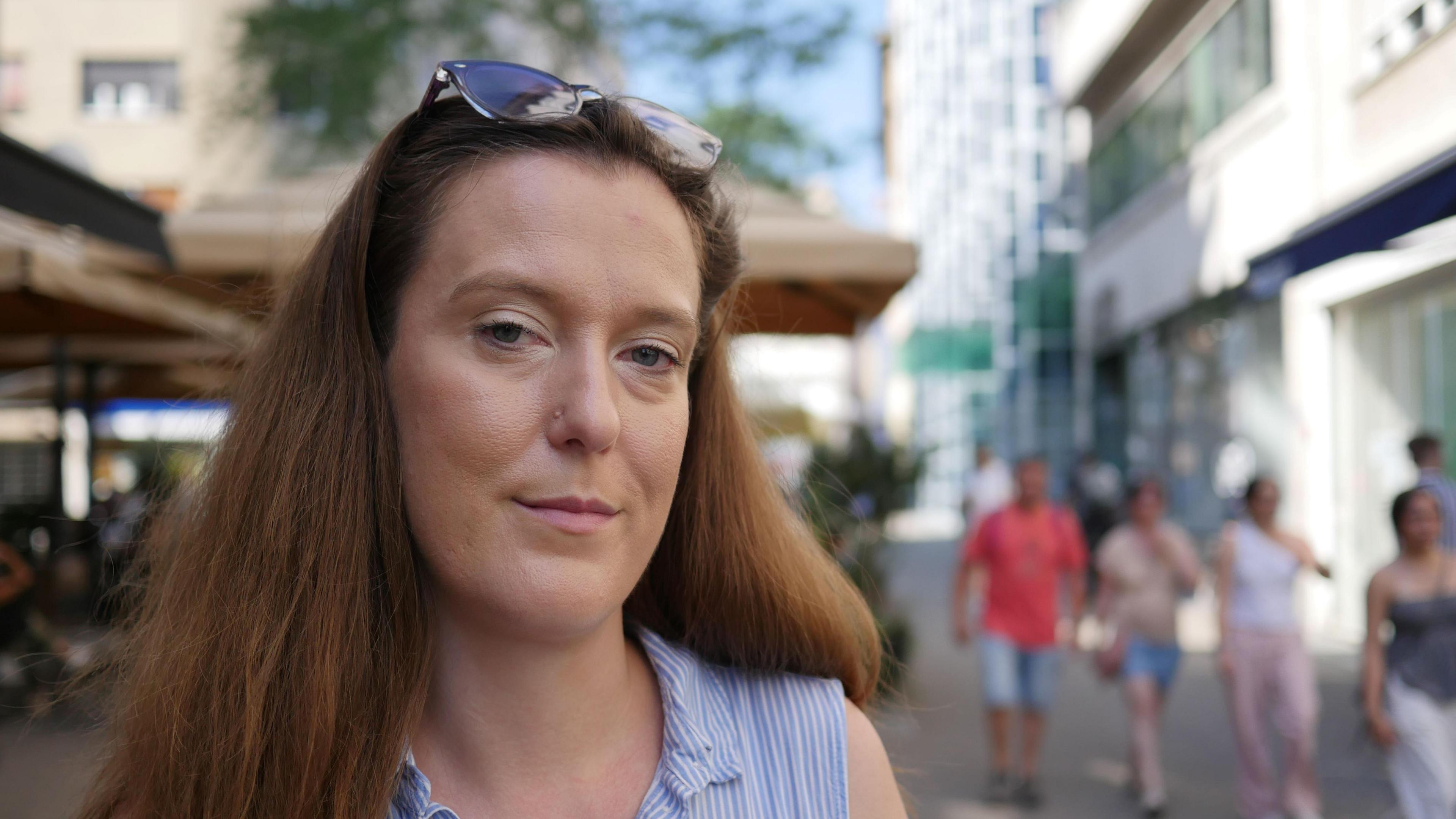Tena Banjeglav looks into the camera. She has long brown hair and is wearing sunglasses on her head. She is standing on the pavement of a quiet street.