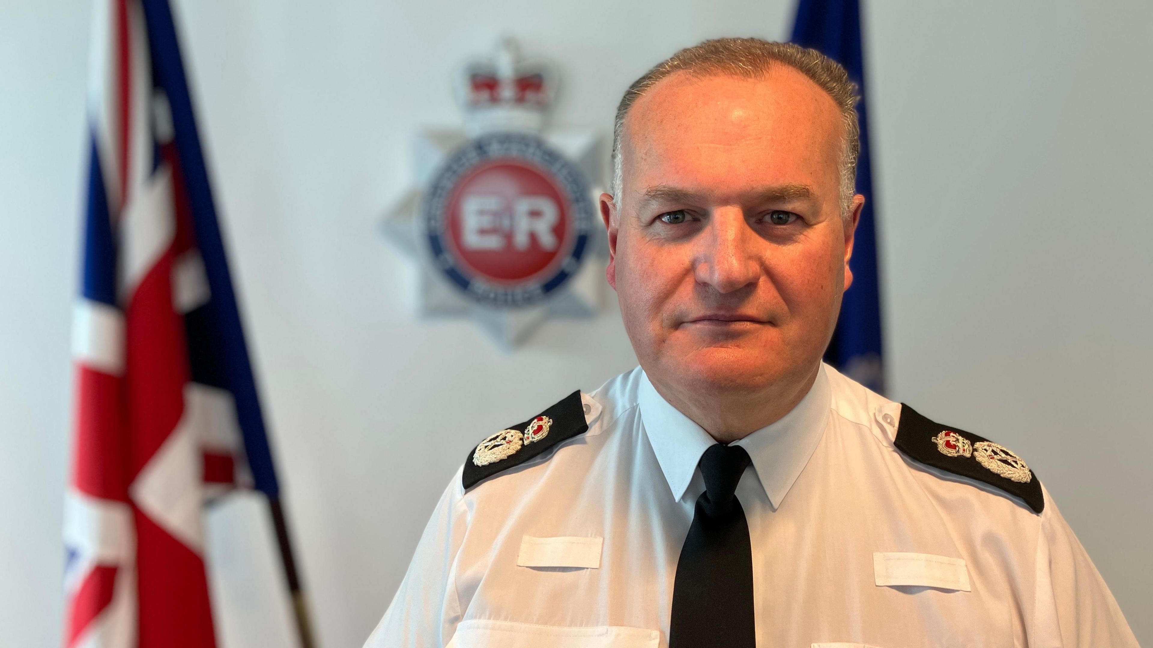 Chief Constable Stephen Watson is starring directly at the camera in his police uniform. The Greater Manchester Police logo is hanging on the wall behind him and there are Union Jack flags on either side.