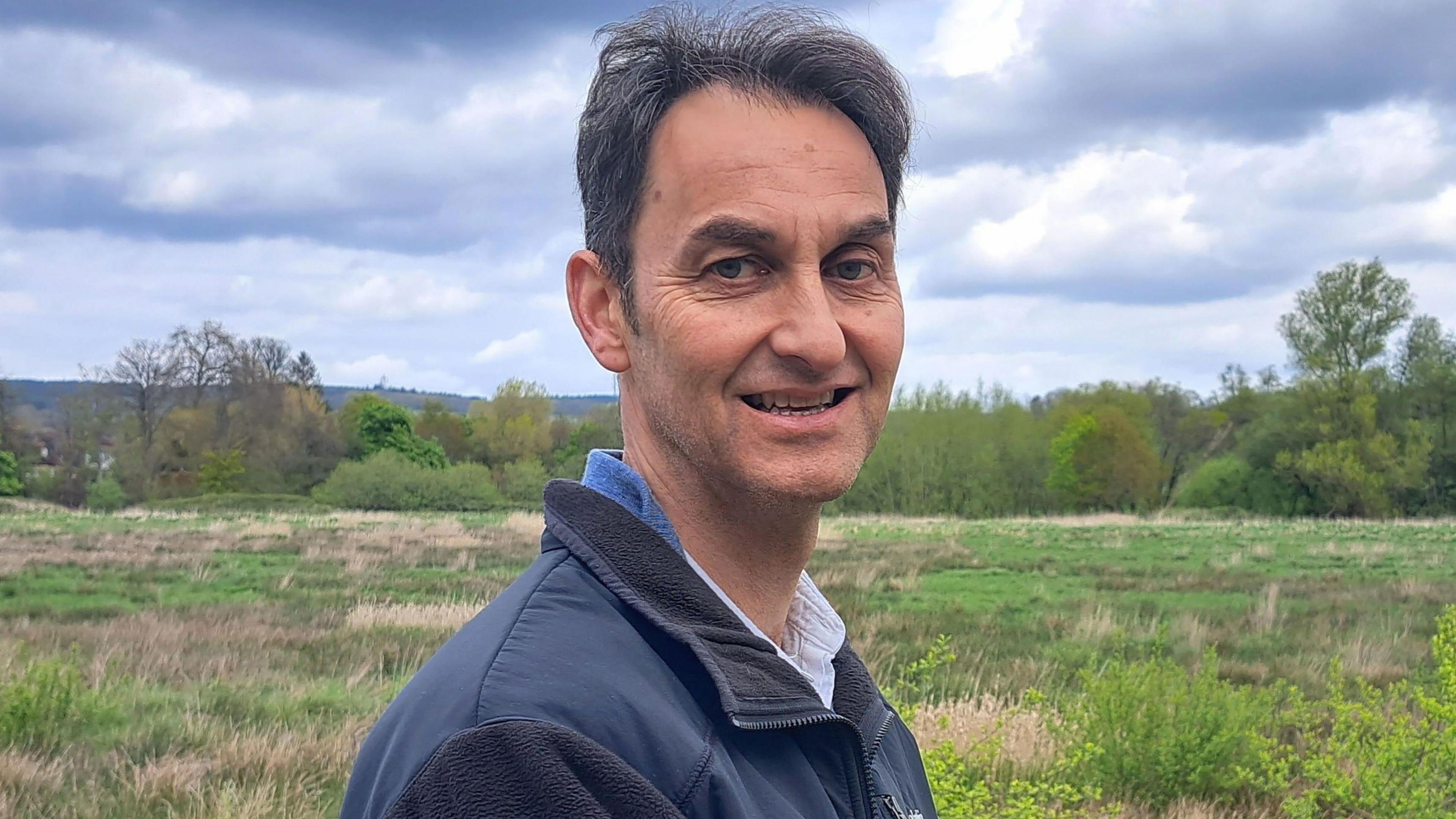 Tim Slaney, a white man in his sixties with short greying dark hair looks at the camera smiling with South Downs National Park greenery in the background