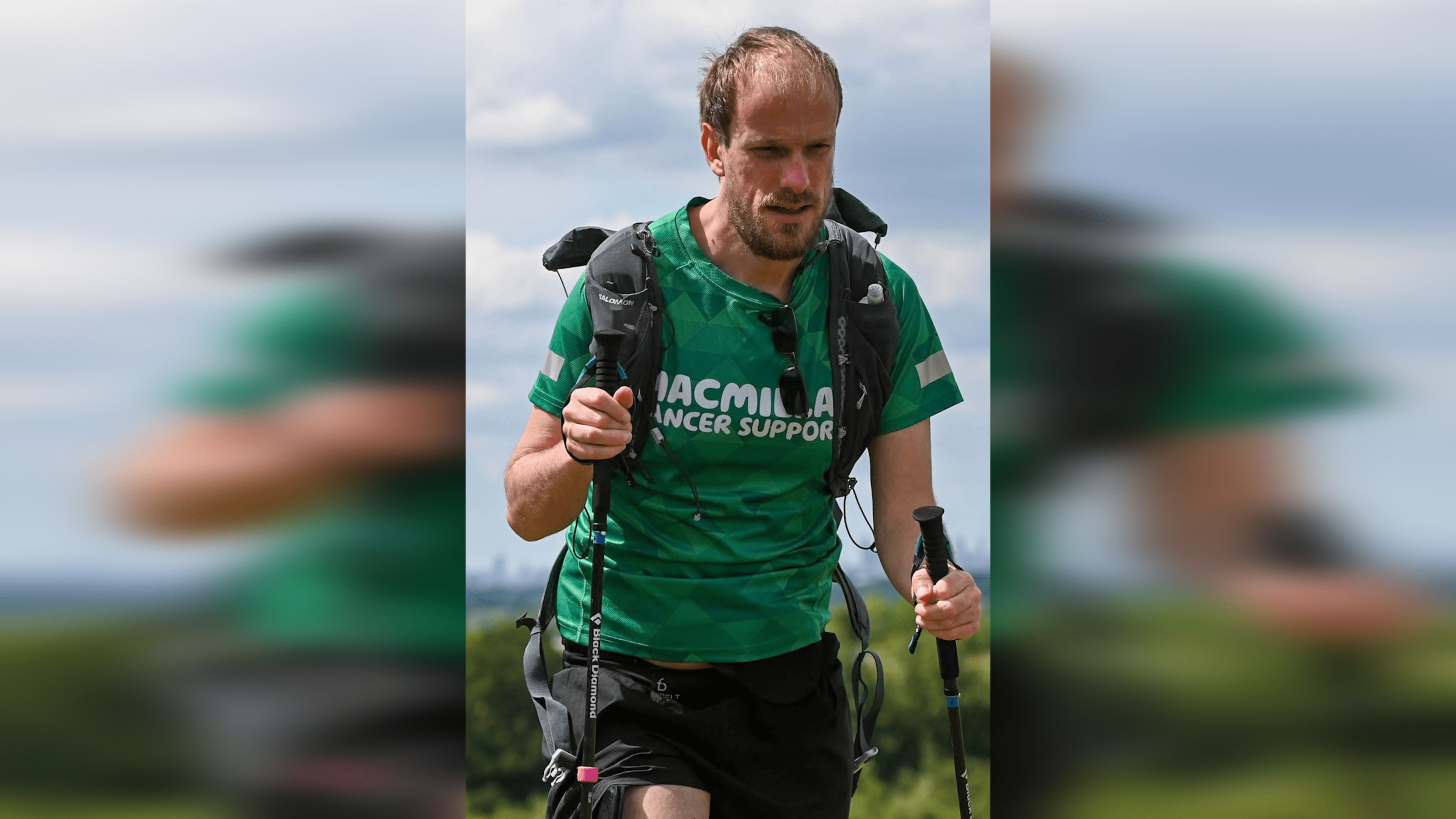 Mr Dye wearing a green T-shirt that reads: Macmillan Cancer Support, carrying walking poles and wearing a black rucksack. He is walking in the countryside.