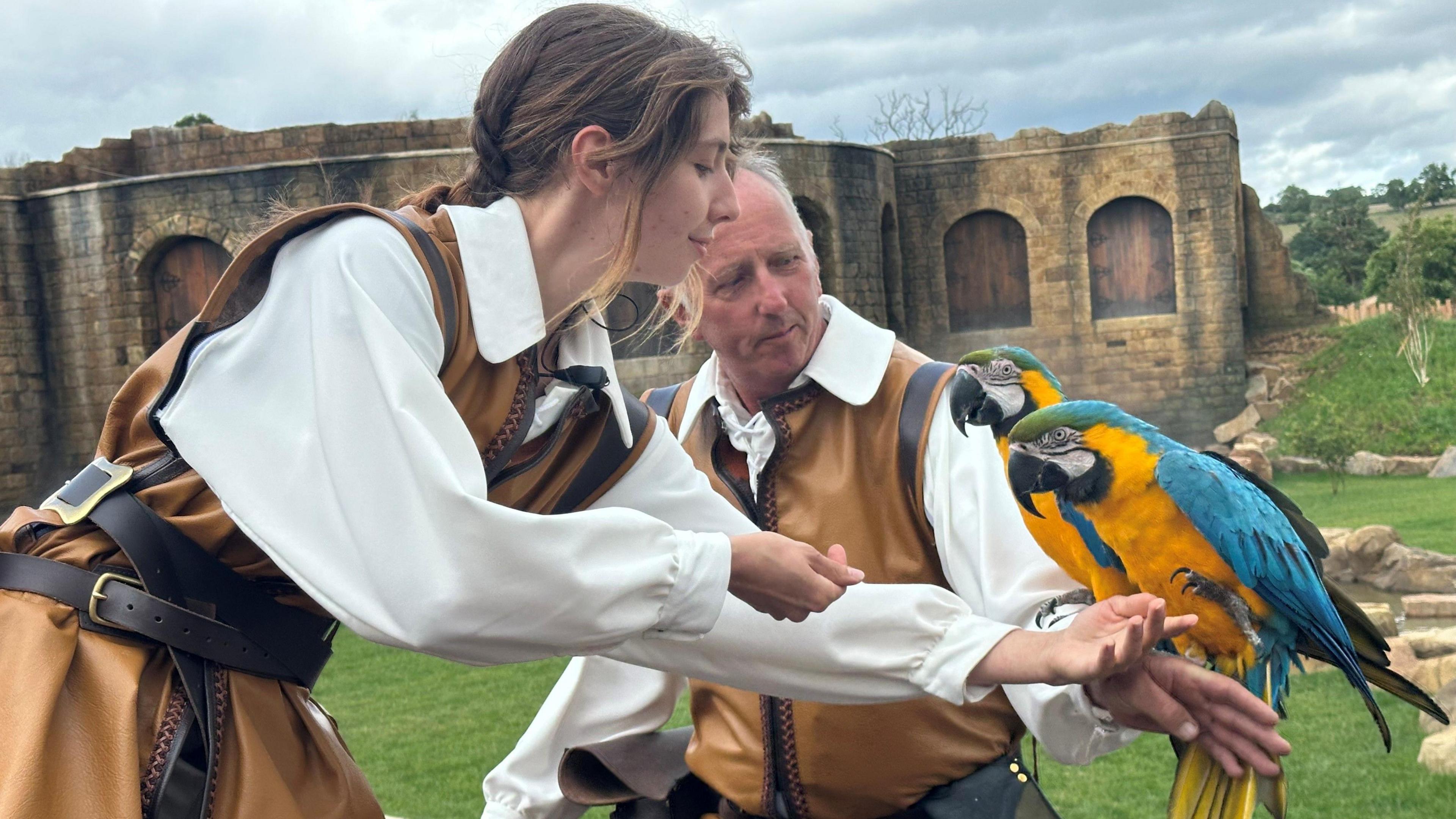 Two keepers, one a man and the other a woman, wearing Tudor-style brown tunics and white shirts, hand a pair of blue and yellow macaws to each other.