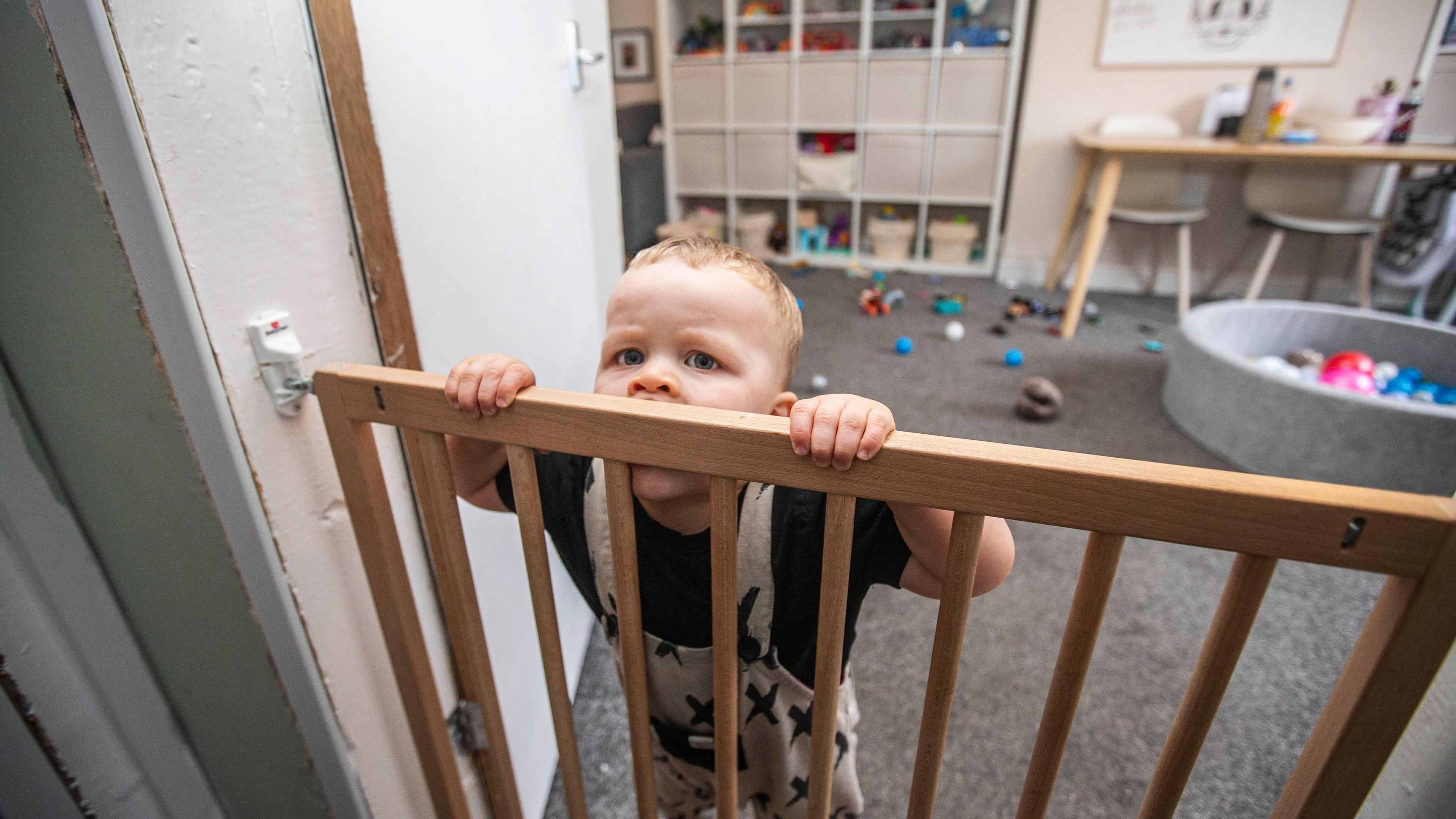 A young boy wearing a black t shirt and black and white dungarees. He is stood at a wooden baby gate, with his hands and mouth around it. Behind him is a play room with toys and a ball pit.