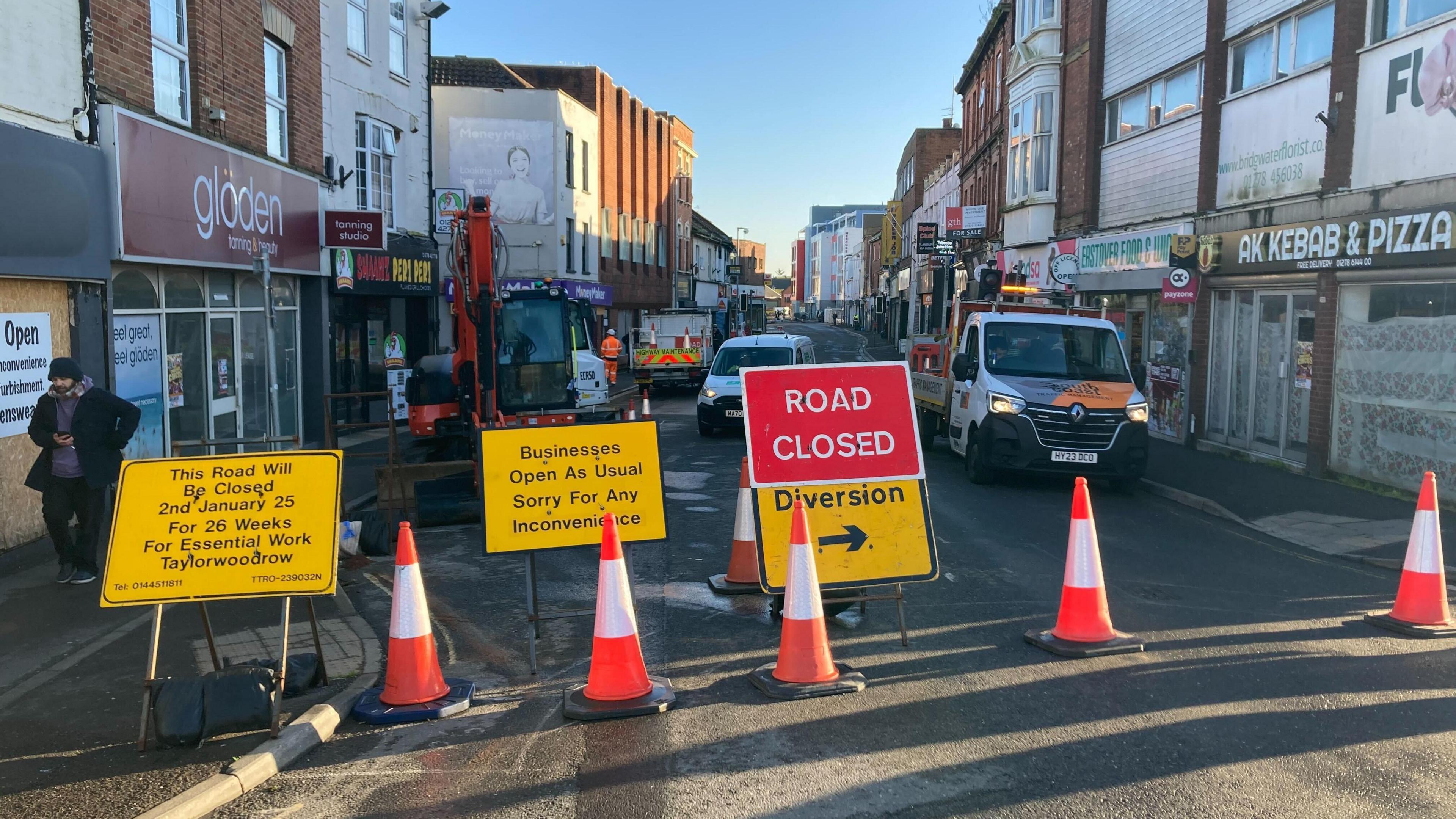 A number of traffic cones block a busy shopping street. There are also four large signs. The first one says "this road will be closed 2nd January 25 for 26 weeks for essential work Taylorwoodrow", while the second says "businesses open as usual, sorry for any inconvenience". A third sign says 'road closed', while the other says 'diversion'. A number of work vans are visible in the background, as well as a digger and a worker in bright orange high-vis. 