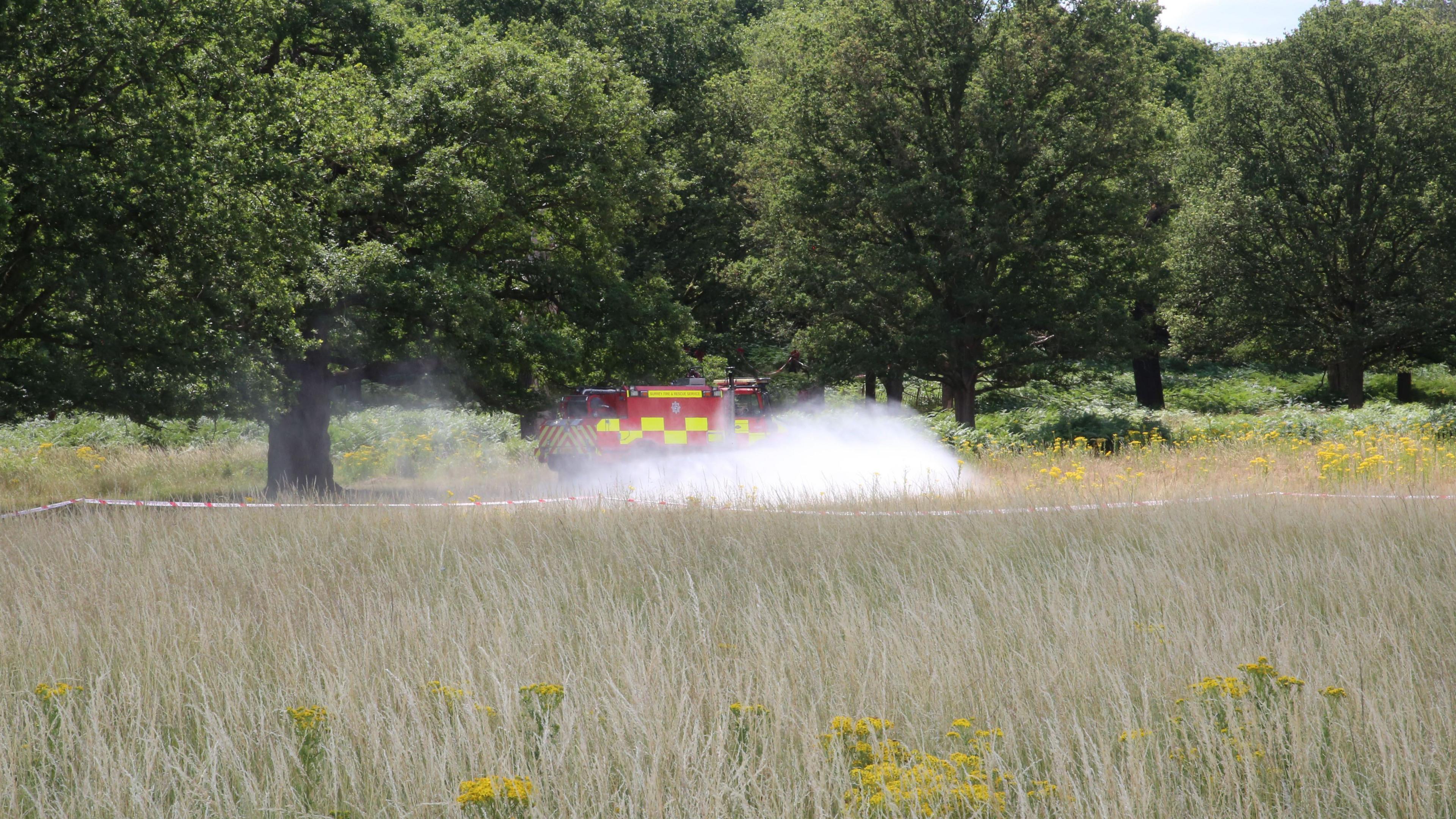Water sprays horizontally from a red fire truck into a field of long grass with tree behind it
