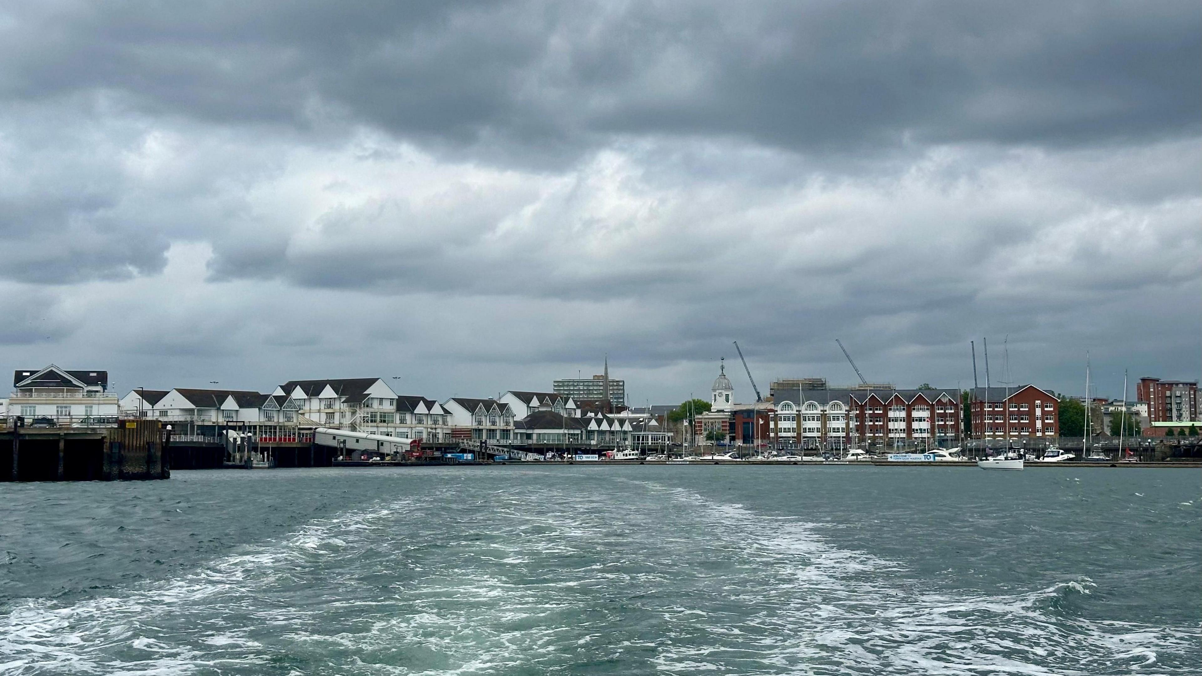 SUNDAY - Looking back from a boat towards Town Quay in Southampton. The boats wake is showing white in Southampton Water, behind you can see the passenger ferry pontoon and the quay with cars parked. In the distance sailing boats are moored in front of white and brick buildings.