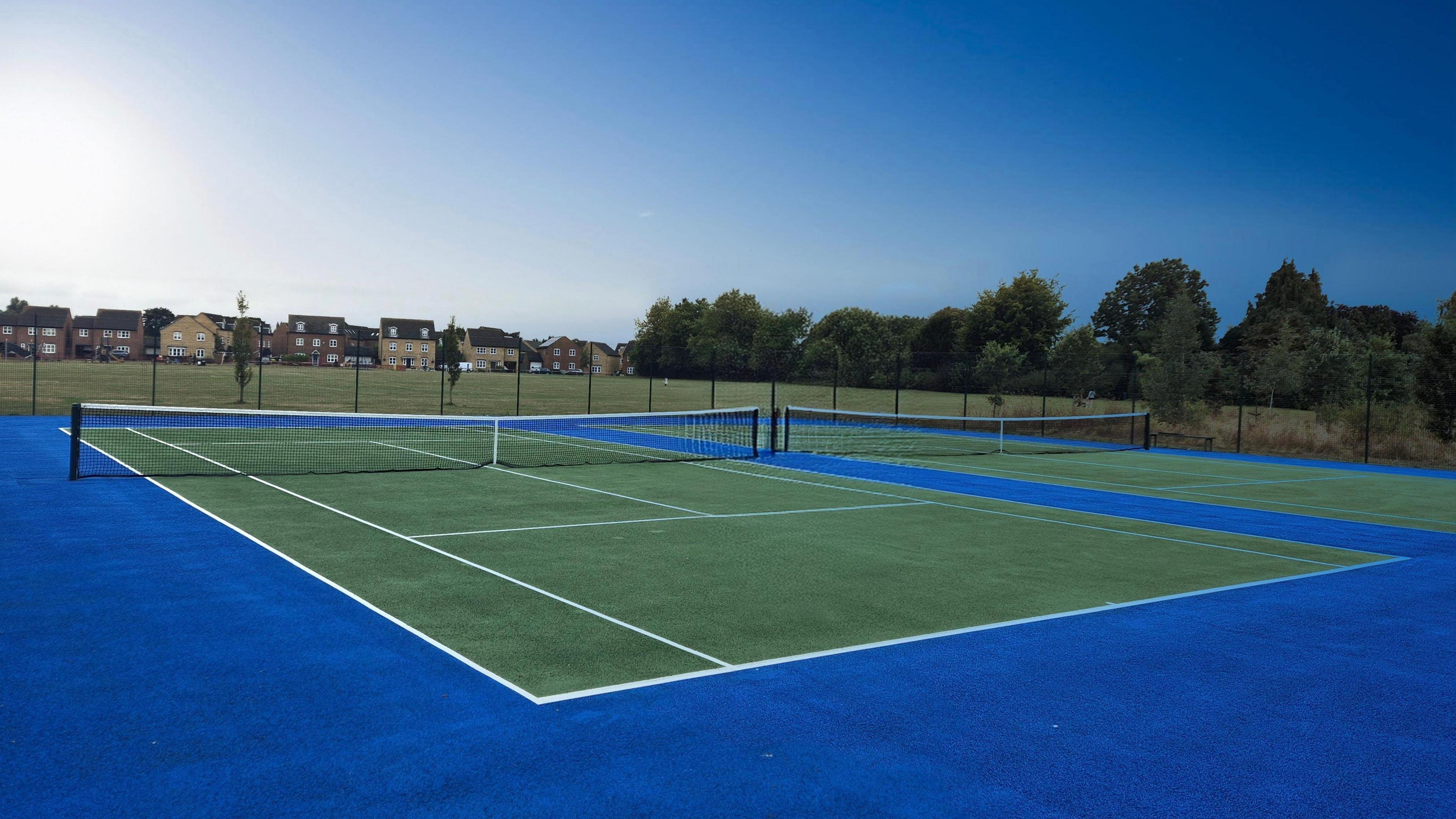 Two hard tennis courts with revamped surfaces. The courts are dark green and have a royal blue perimeter. Outside of the fenced courts is a public park with grass, surrounded by trees and houses. It is a sunny day.