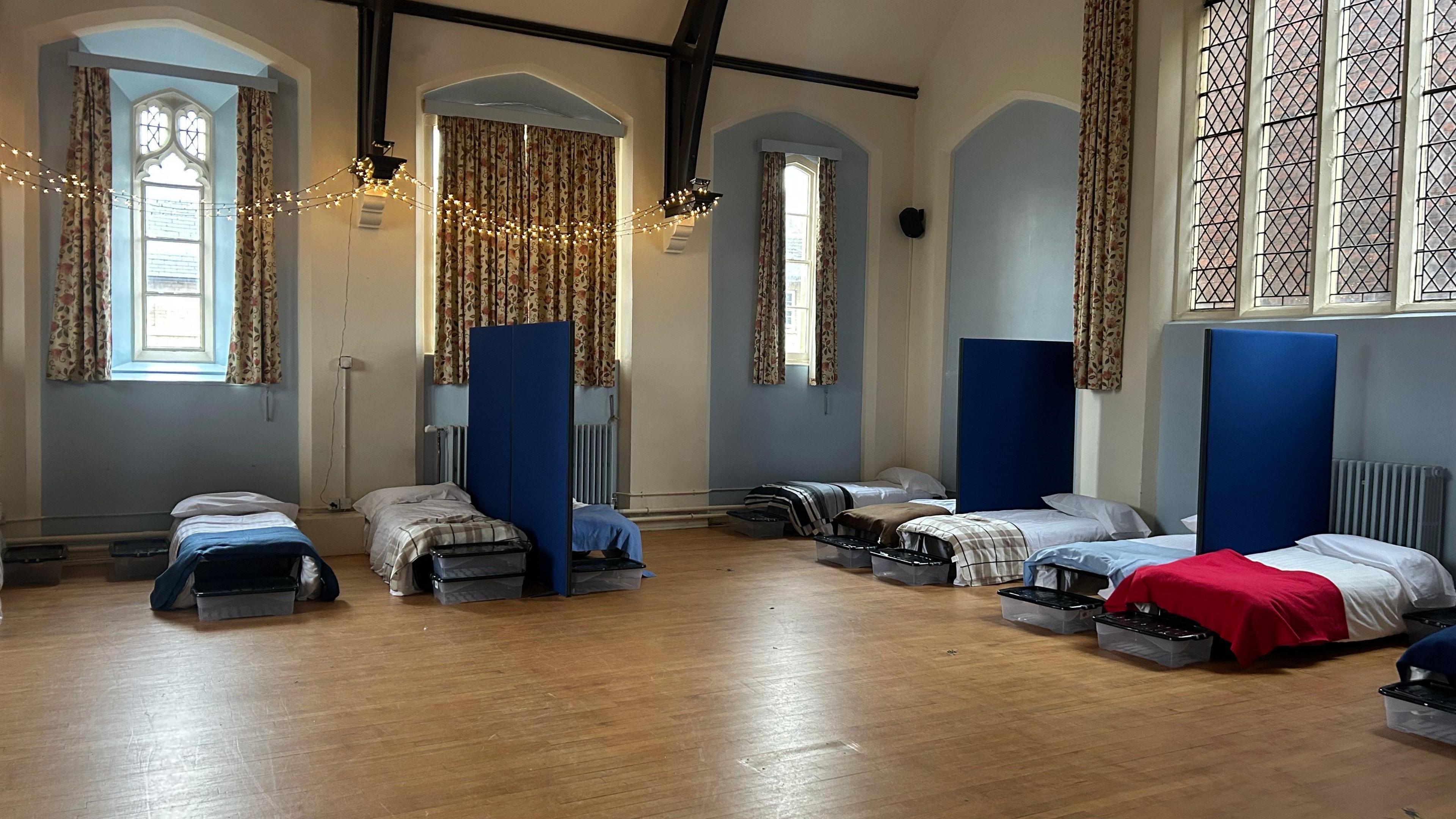 A room, with Christmas lights put up on one wall, with nine beds, with bedding in them, and blue partitions. The floor is wooden, there are large curtains at a number of windows and the beds all have different bedding on. Boxes are at the end of the bed. 