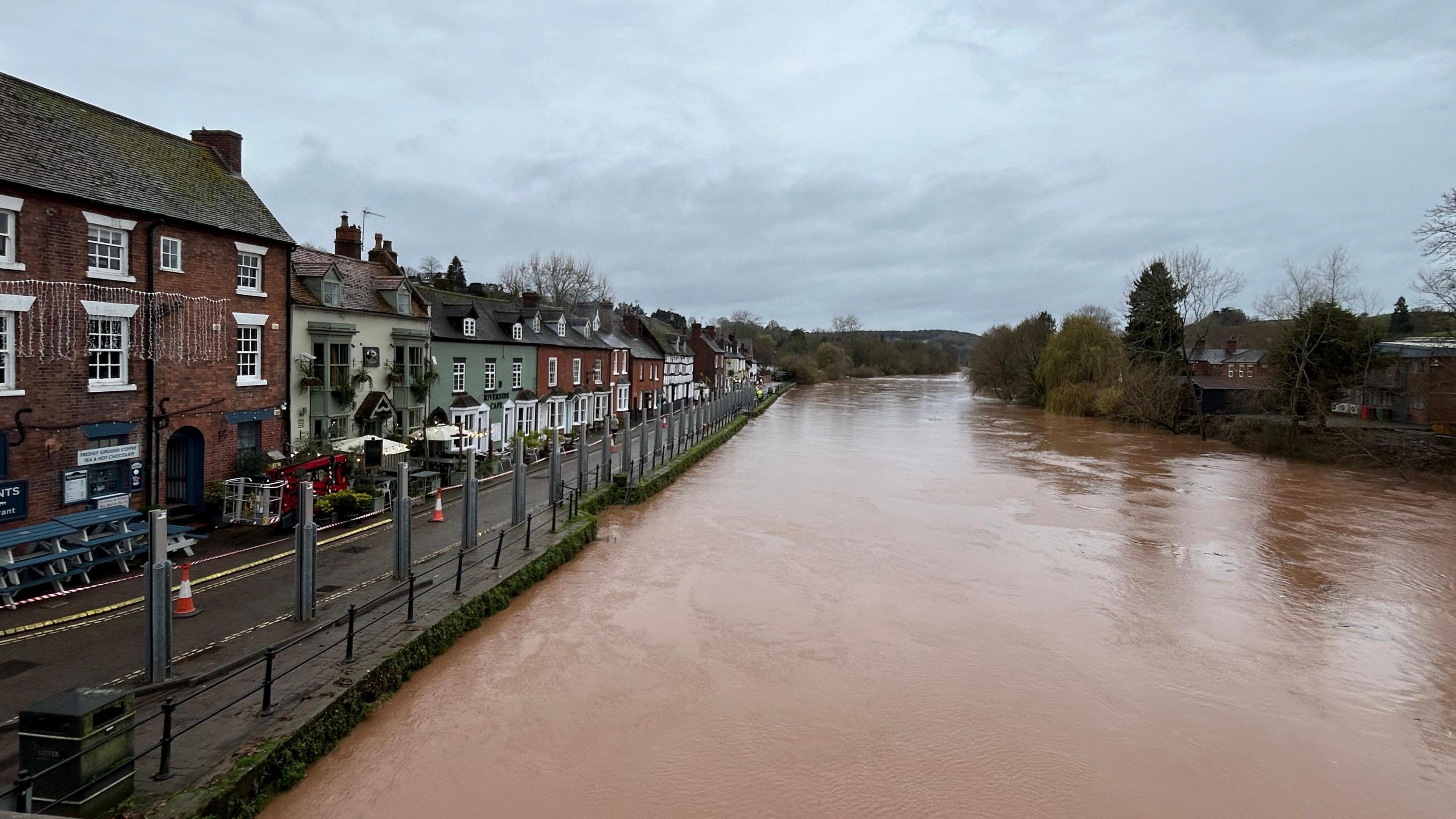 a river with brown water. alongside the river are rows of houses with silver metal poles, flood defence barriers, aligned along the side of the river.