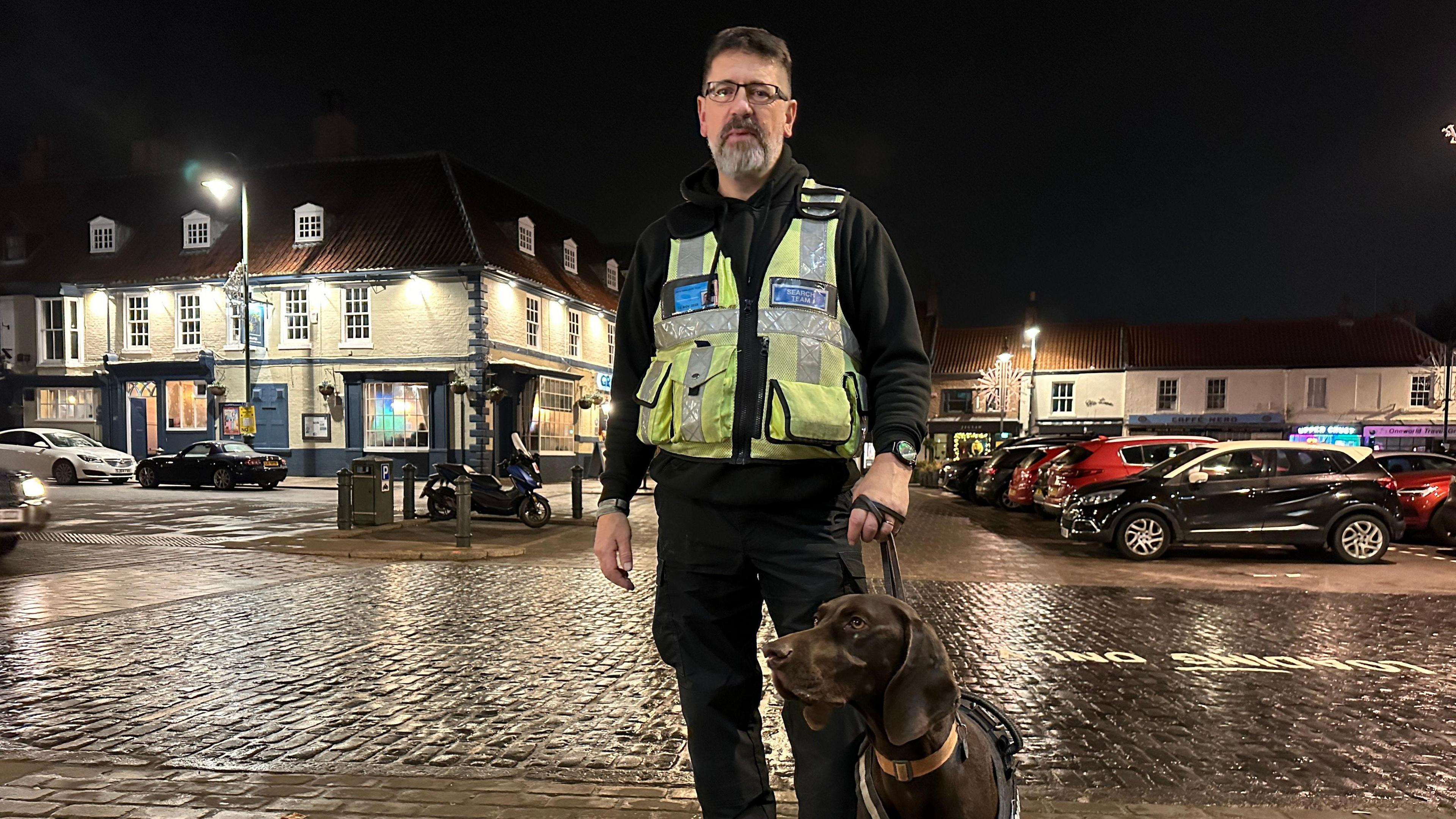 A police dog handler, wearing a black uniform and a yellow hi-vis vest, holds a brown English pointer dog by the lead. They are standing in the cobbled Saturday Market, Beverley, at night, with bars in the background.