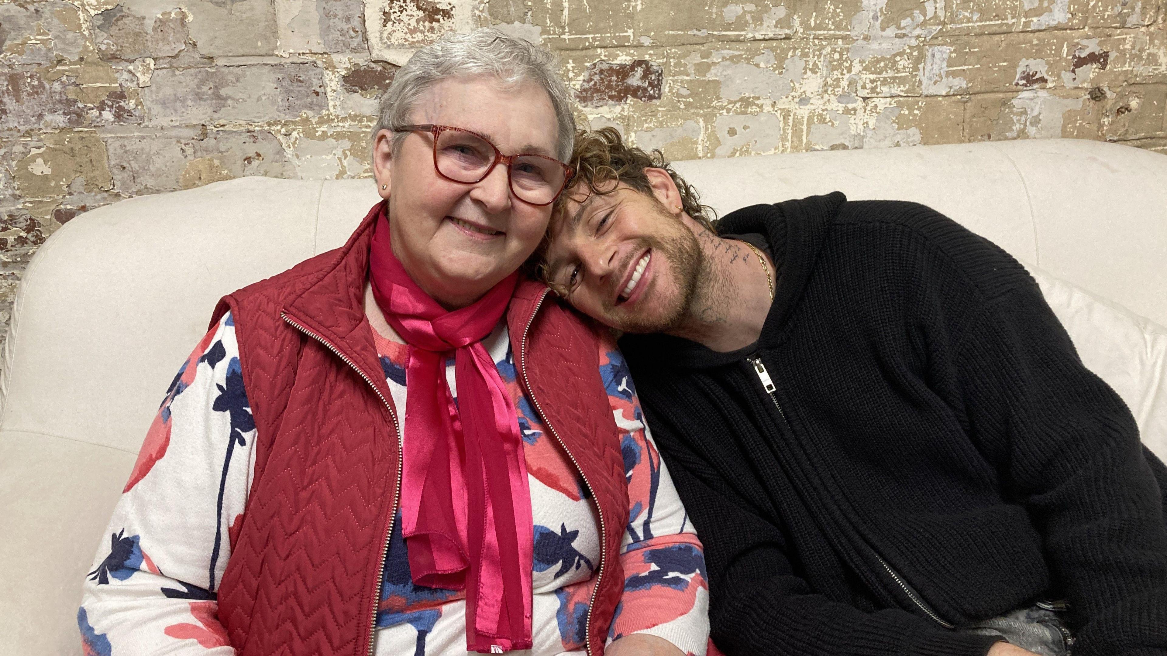 Tom Grennan, who is wearing a black hoodie, leaning his head on his gran's shoulder. She has short grey hair with red glasses, and a red jacket with red scarf.