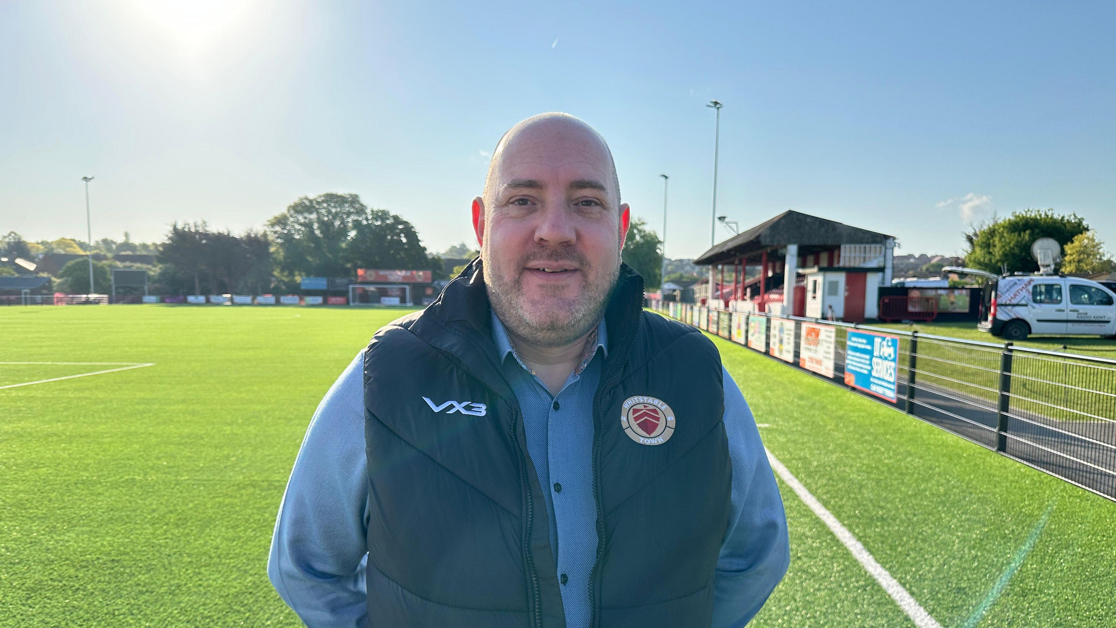 Chairman Steve Clayton looks at the camera and is wearing a blue shirt with a Whitstable gilet over the top. He is standing on the football pitch.