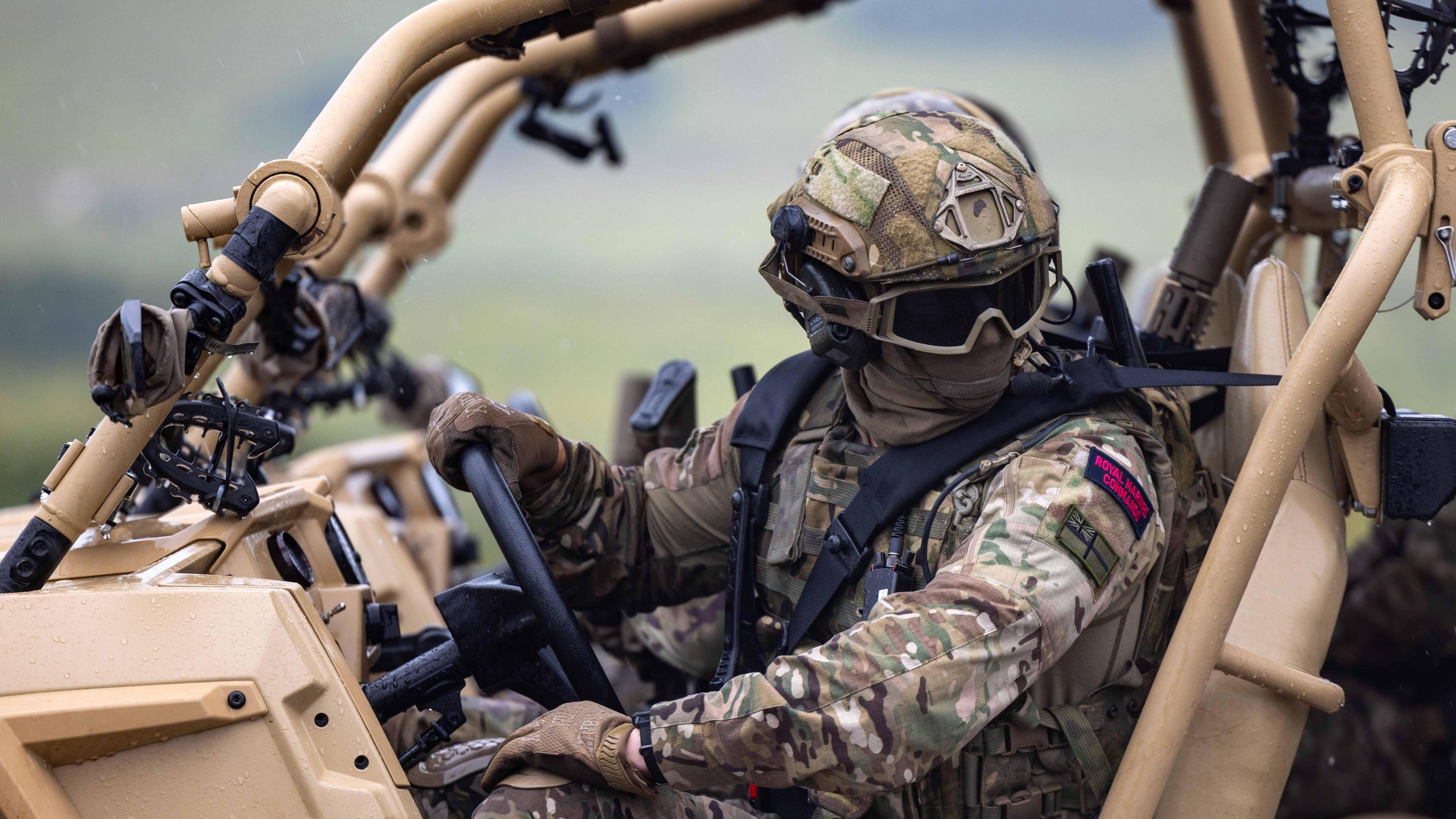 A British Marine Commando sits in an armoured vehicle. His identity is obscured by tactical glasses and a facemask.