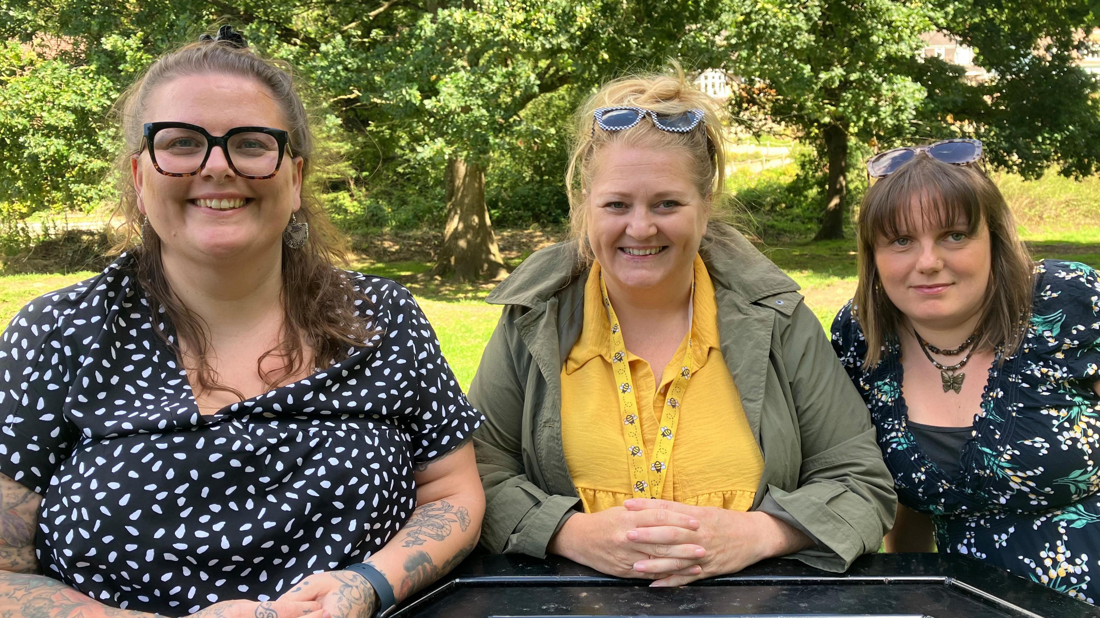 Three women sitting side-by-side on a park picnic bench. They are smiling at the camera and there are trees in dappled sunshine in the background.