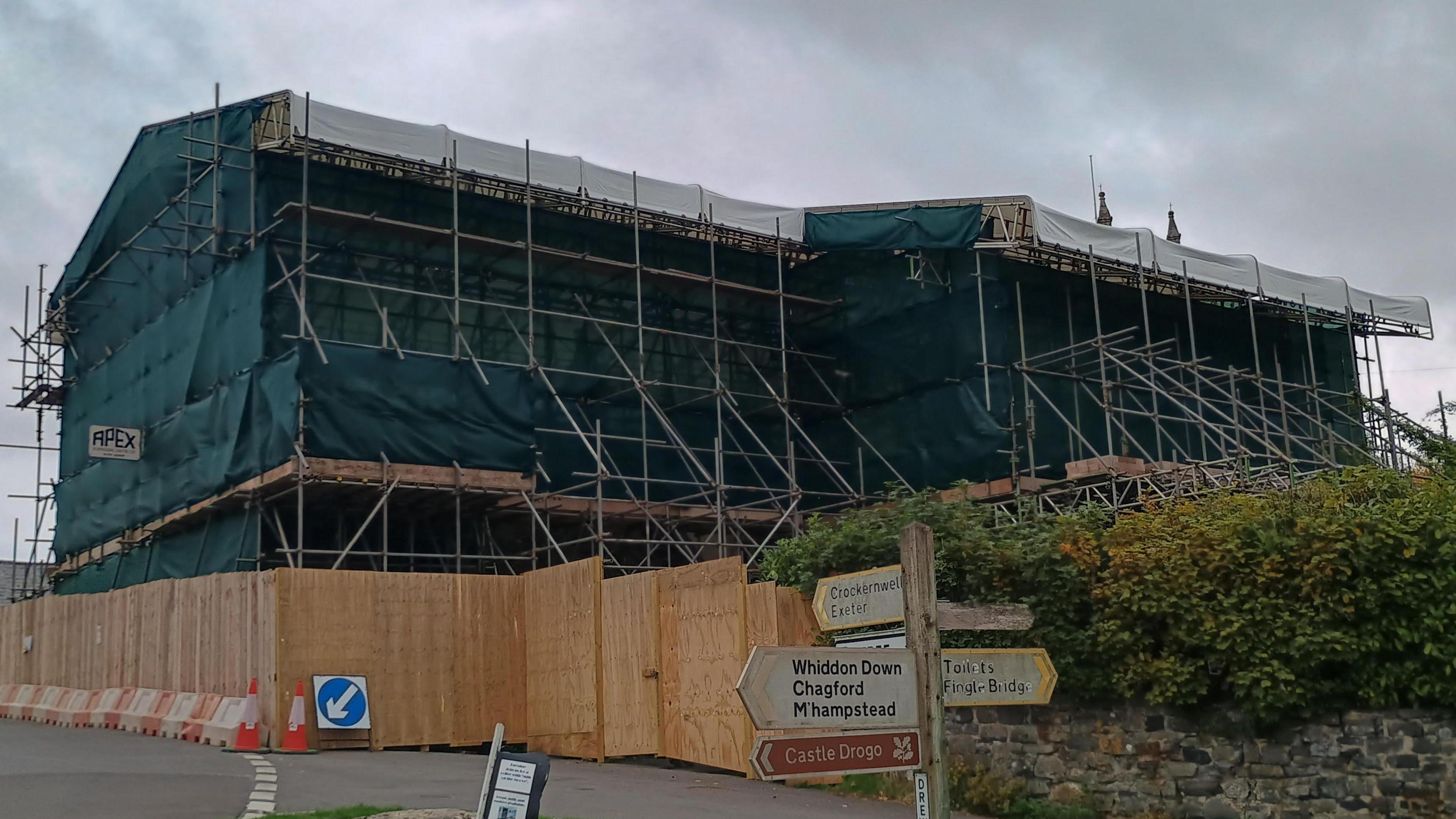 A row of cottages completely covered in scaffolding and tarpaulin. There is wooden fencing around the buildings. There is also a road sign and cones.