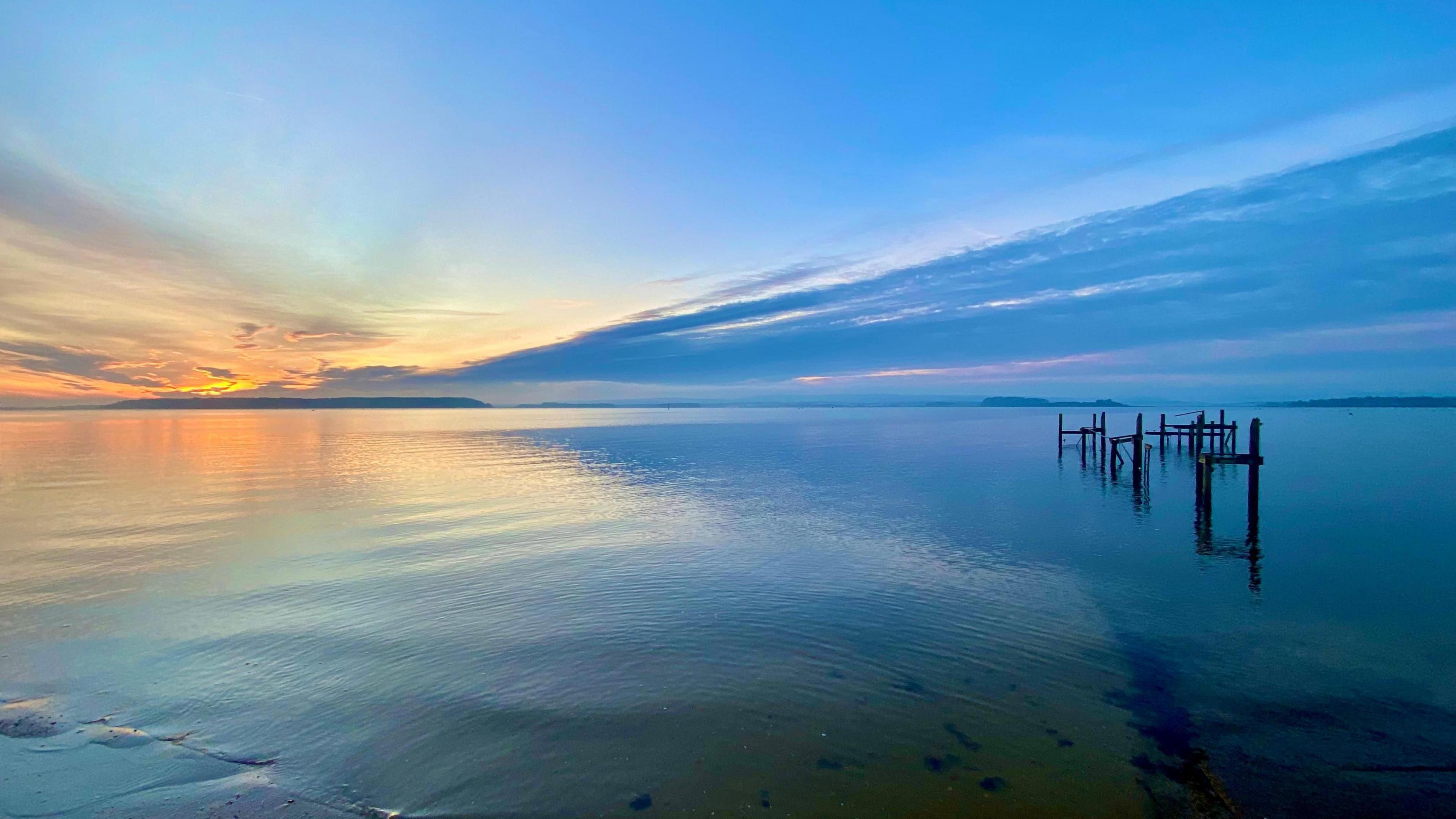 Most of the shot shows a wide expanse of water with wooden groynes on the right-hand side. Both the water and the sky are a very vivid blue colour that darkens from left to right. The first rays of the sun also create a golden glow on the horizon on the left-hand side of the picture.