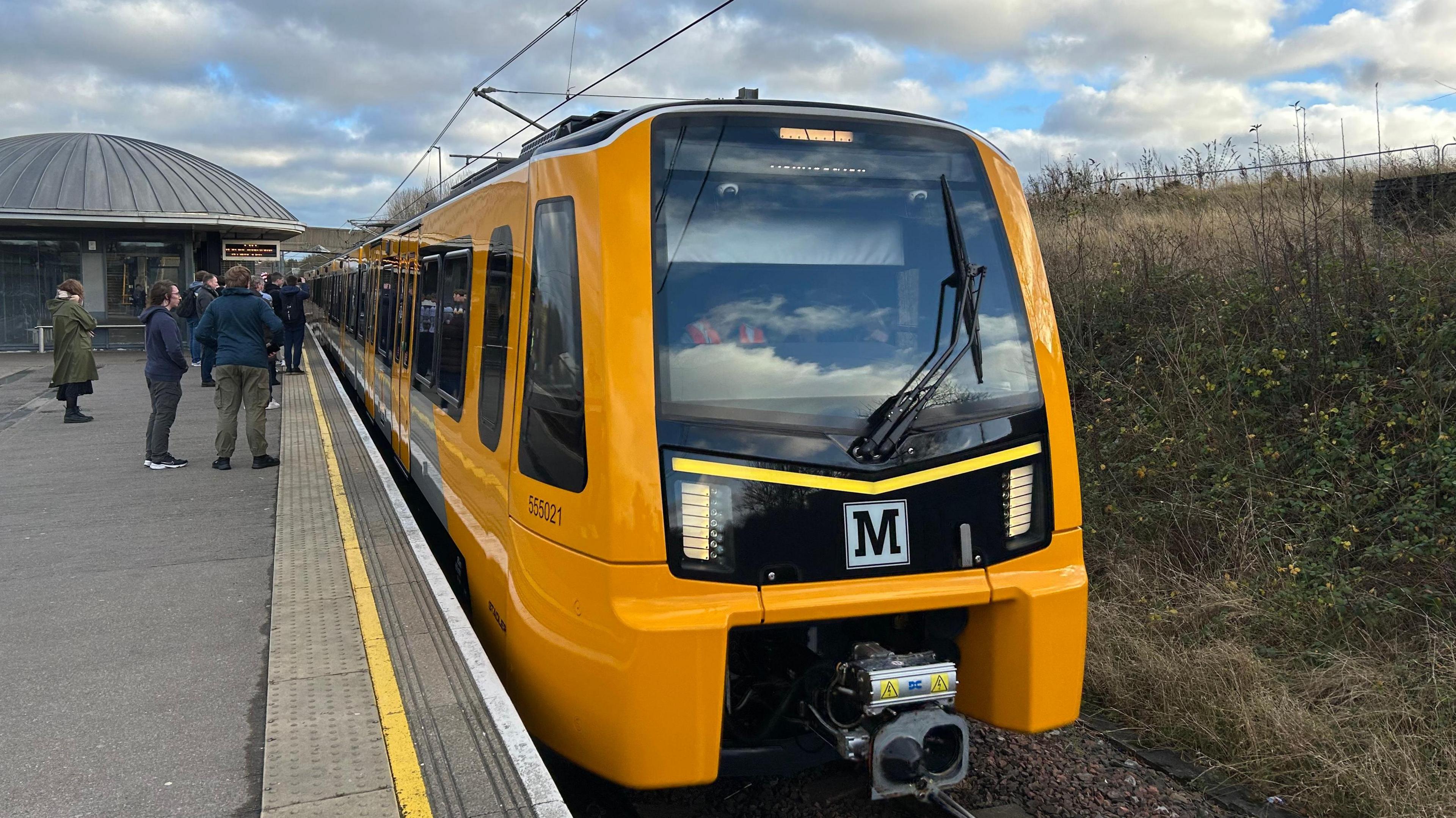 A new model of metro train at Pelaw station. The carriage is yellow with the 'M' metro logo on the front. People are waiting to get on the train.