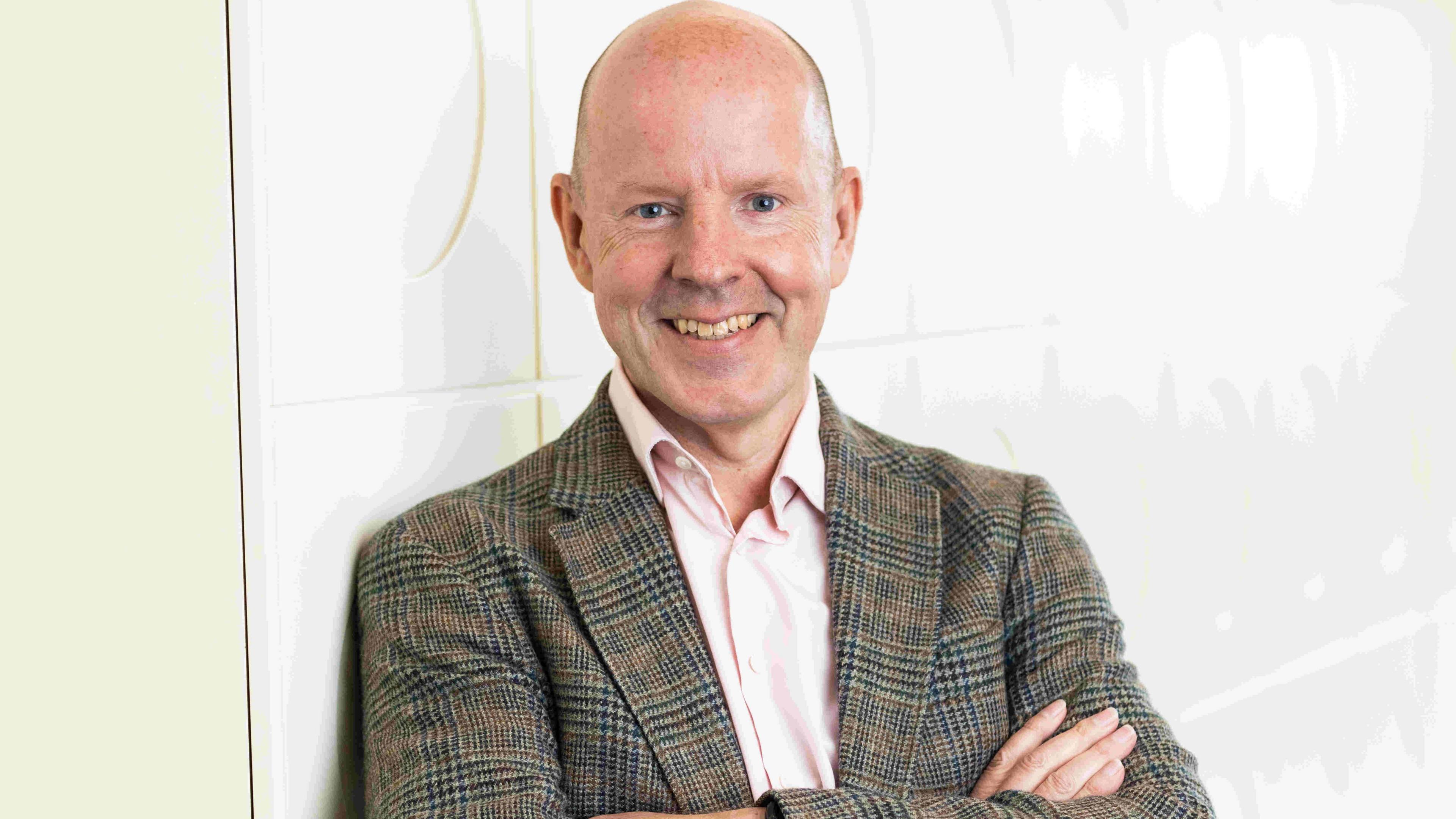 Mark Preece stands facing the camera with his arms crossed, wearing a grey and brown tweed jacket and a pink shirt 