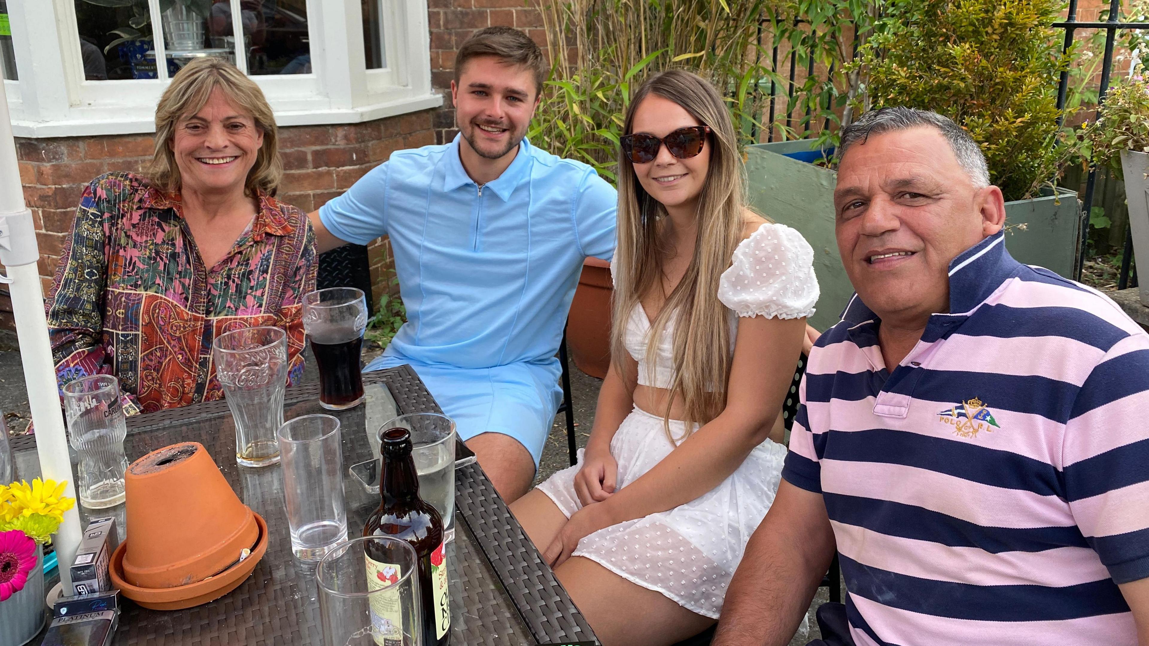 There are four people sitting around a table outside the pub. Jackie Lovell is wearing a colourful shirt sitting next to Dan who is wearing a blue shirt and blue shorts. Glasses are scattered on the table along with bottles of cider and Diet Coke. Dan is sitting next to Georgie who is wearing a white top and skirt and sunglasses. She sits next to David who is wearing a pink and blue striped shirt