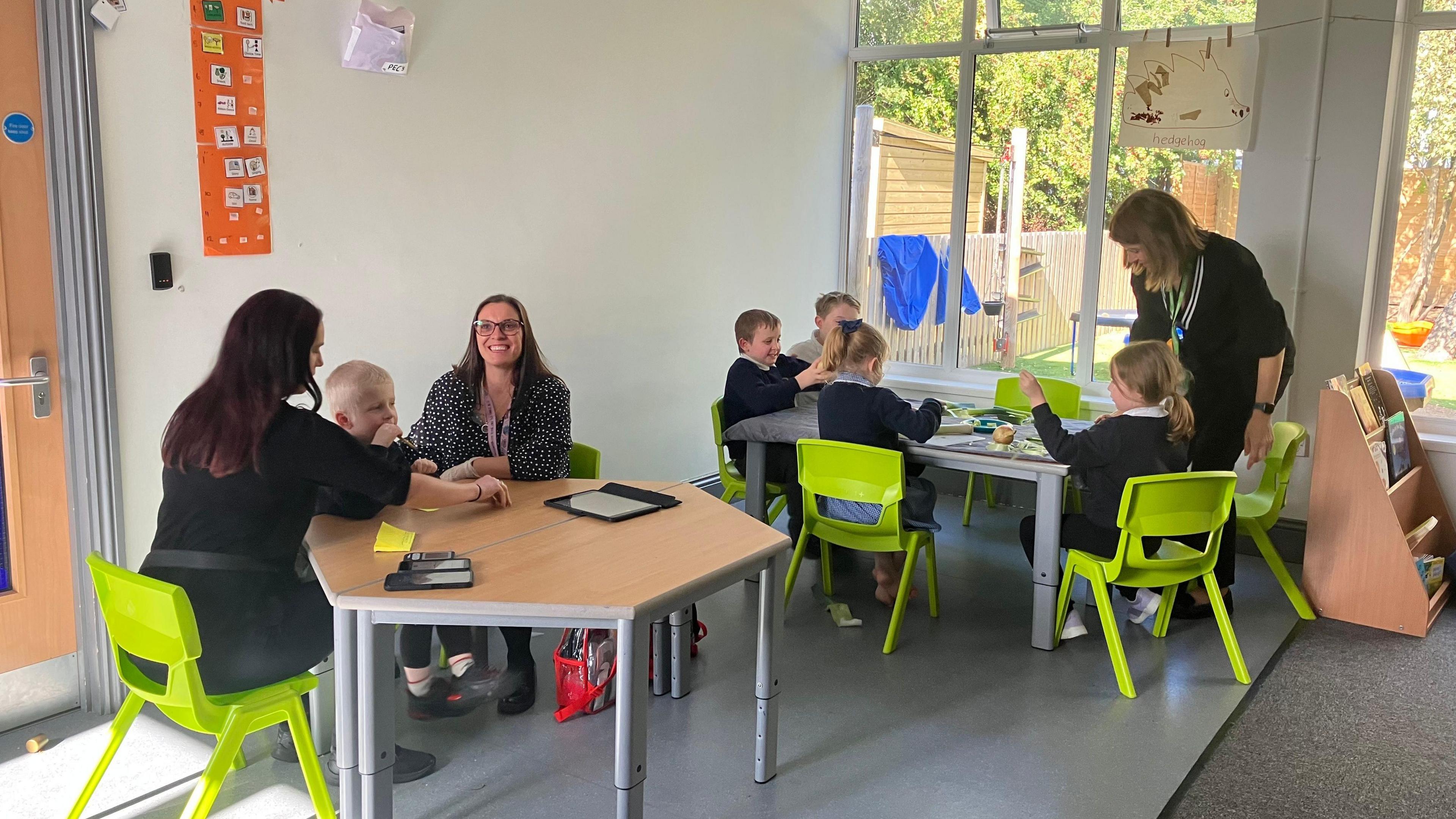 Five pupils aged below six, plus three staff sit across two tables with green chairs in a classroom setting. One table is working with technology, and the other table is chopping vegetables. 