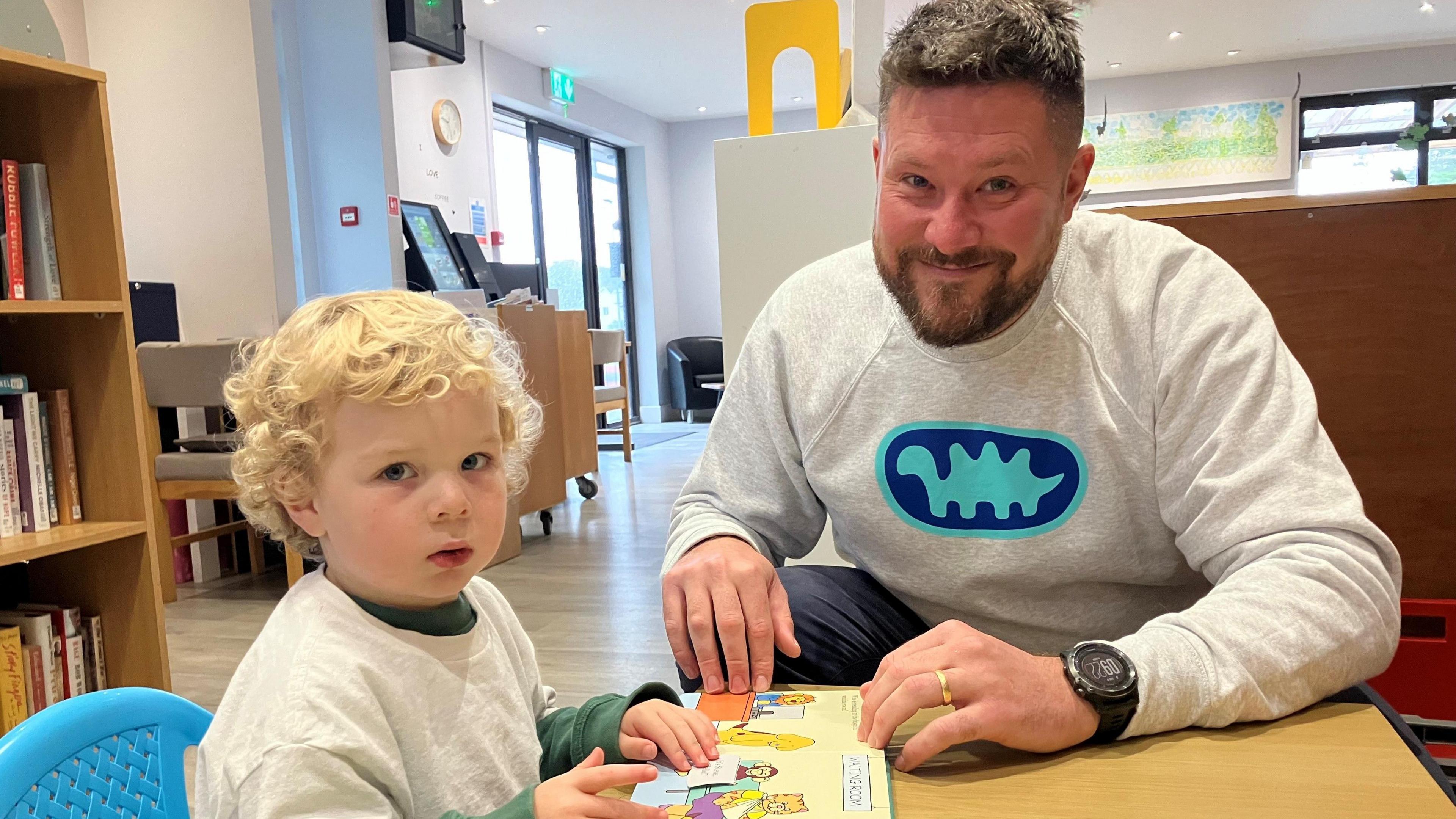 Young boy looking very suspicious at the camera while reading with his dad at Par Track Library 