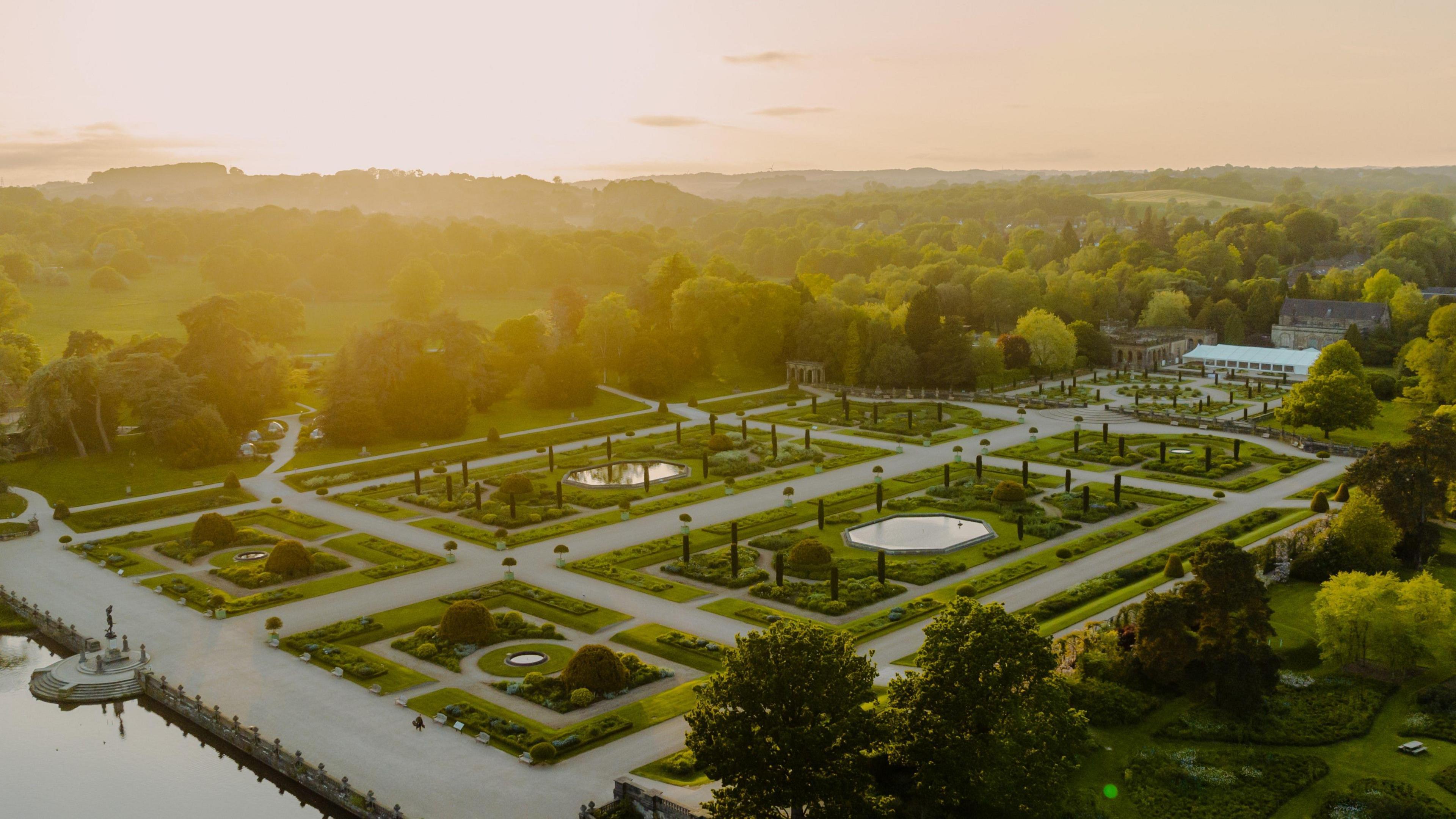 An aerial image of the Trentham Estate gardens with the sun setting on the horizon behind the estate. The gardens are divided into rectangular and square sections featuring trees and other greenery, and are split off by a series of large pathways. The gardens are surrounded by woodland