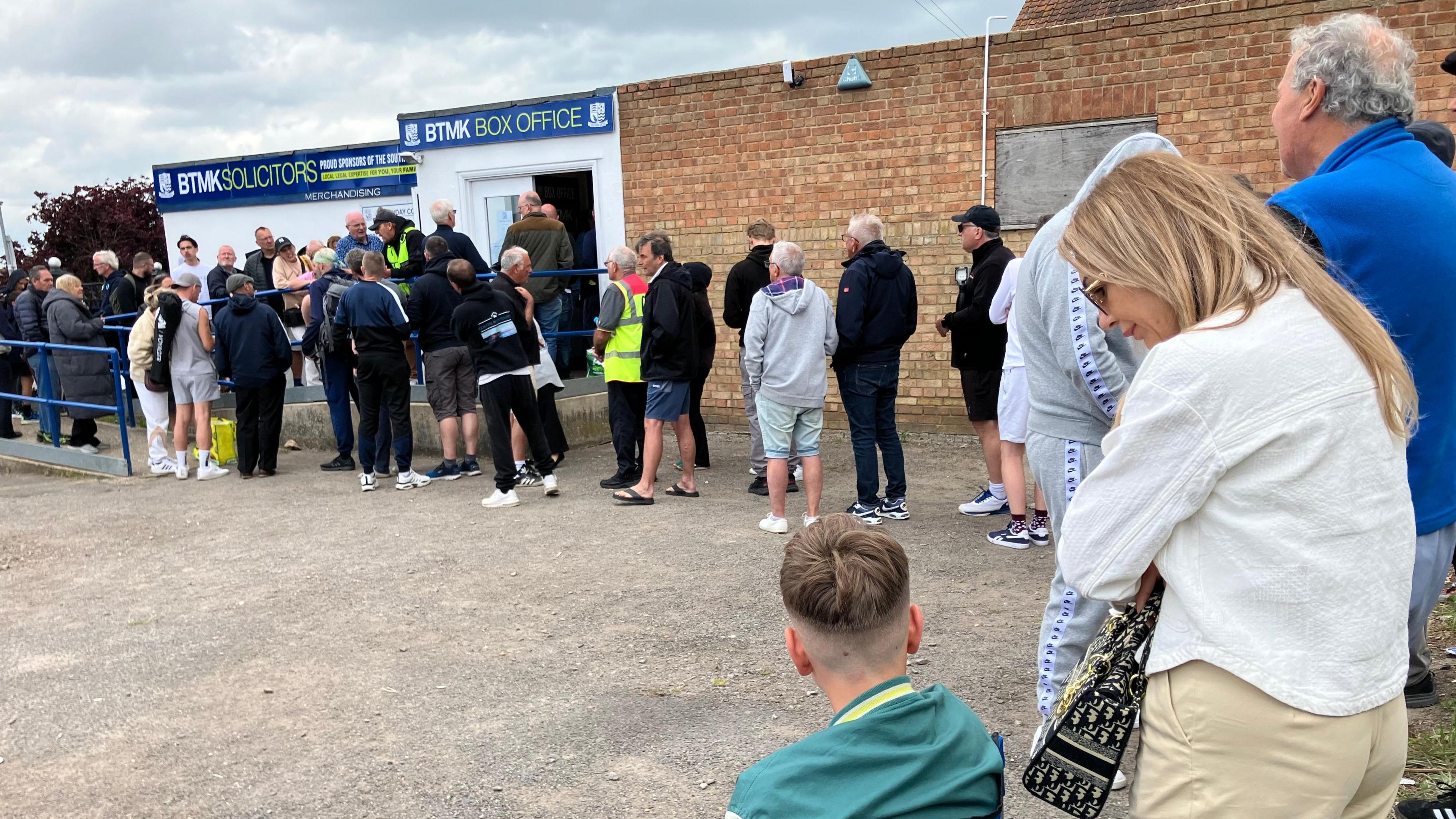 The queue of supporters seen from the car park. It features lots of people winding their way to the ticket office building. In the foreground a woman is looking at a younger man who is sitting on a camping chair.