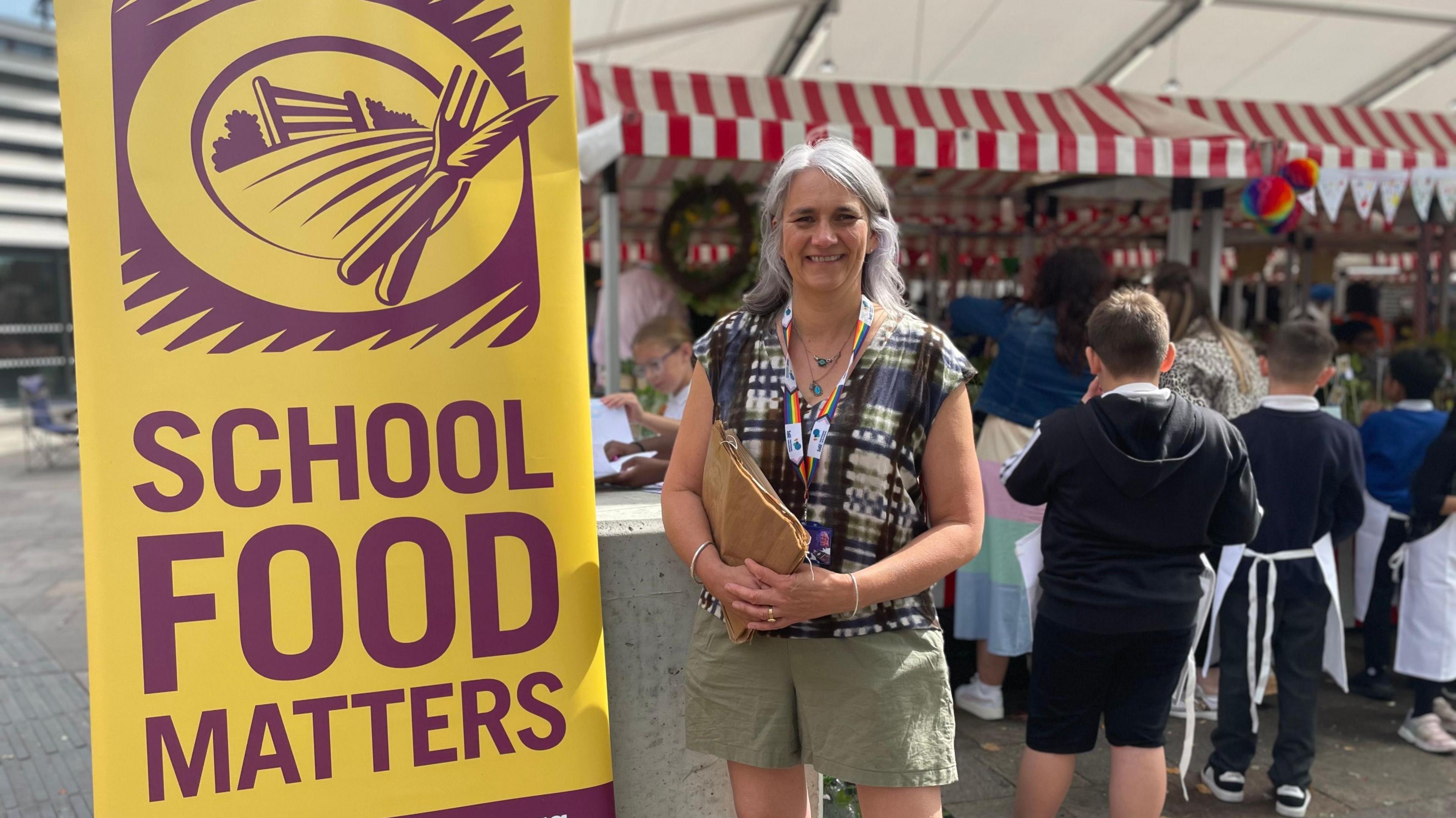 Lisa Didier stood next to a yellow School Food Matters poster. She is holding brown paper bags and is smiling. She is wearing a colourful top and lanyard.