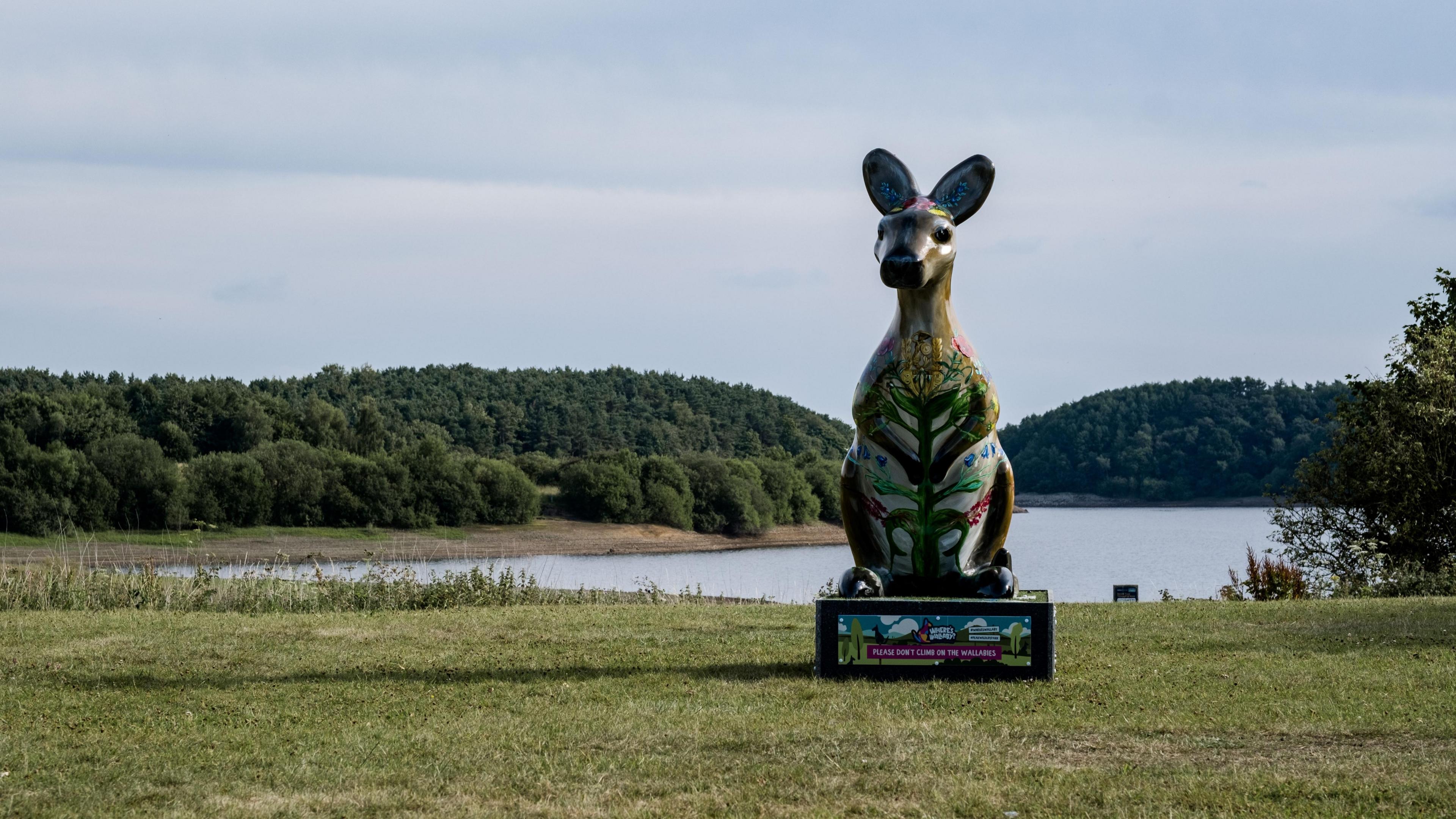 A wallaby sculpture located by a lake, on a patch of grass. There are trees in the distance. The wallaby sculpture is colourfully decorated.