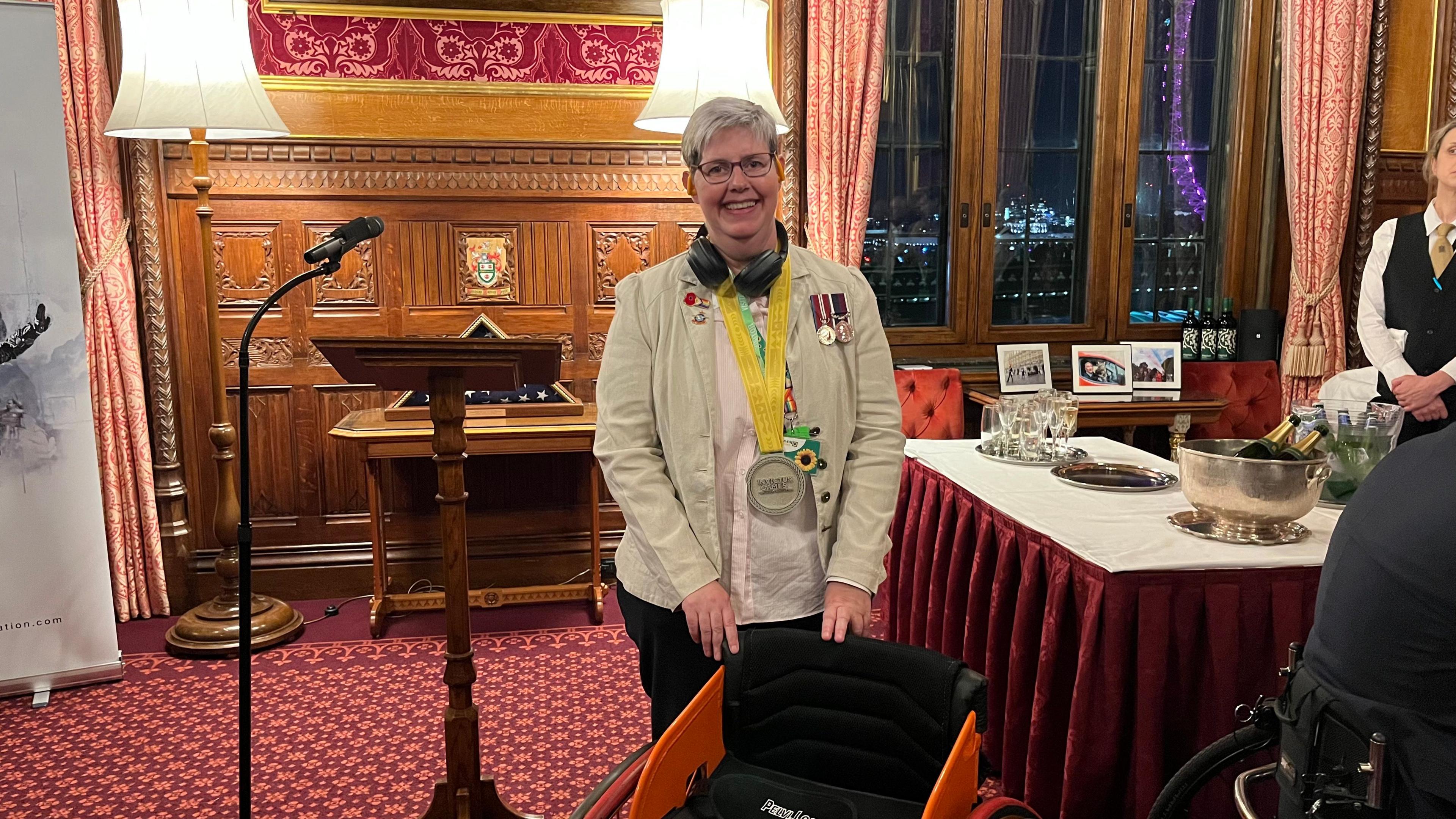 Sara Howarth wearing a light-coloured jacket with multiple medals standing in a wood-panelled room. She is wearing an Invictus Games medal which hangs from a yellow and green ribbon around her neck. In front of her is an orange wheelchair.