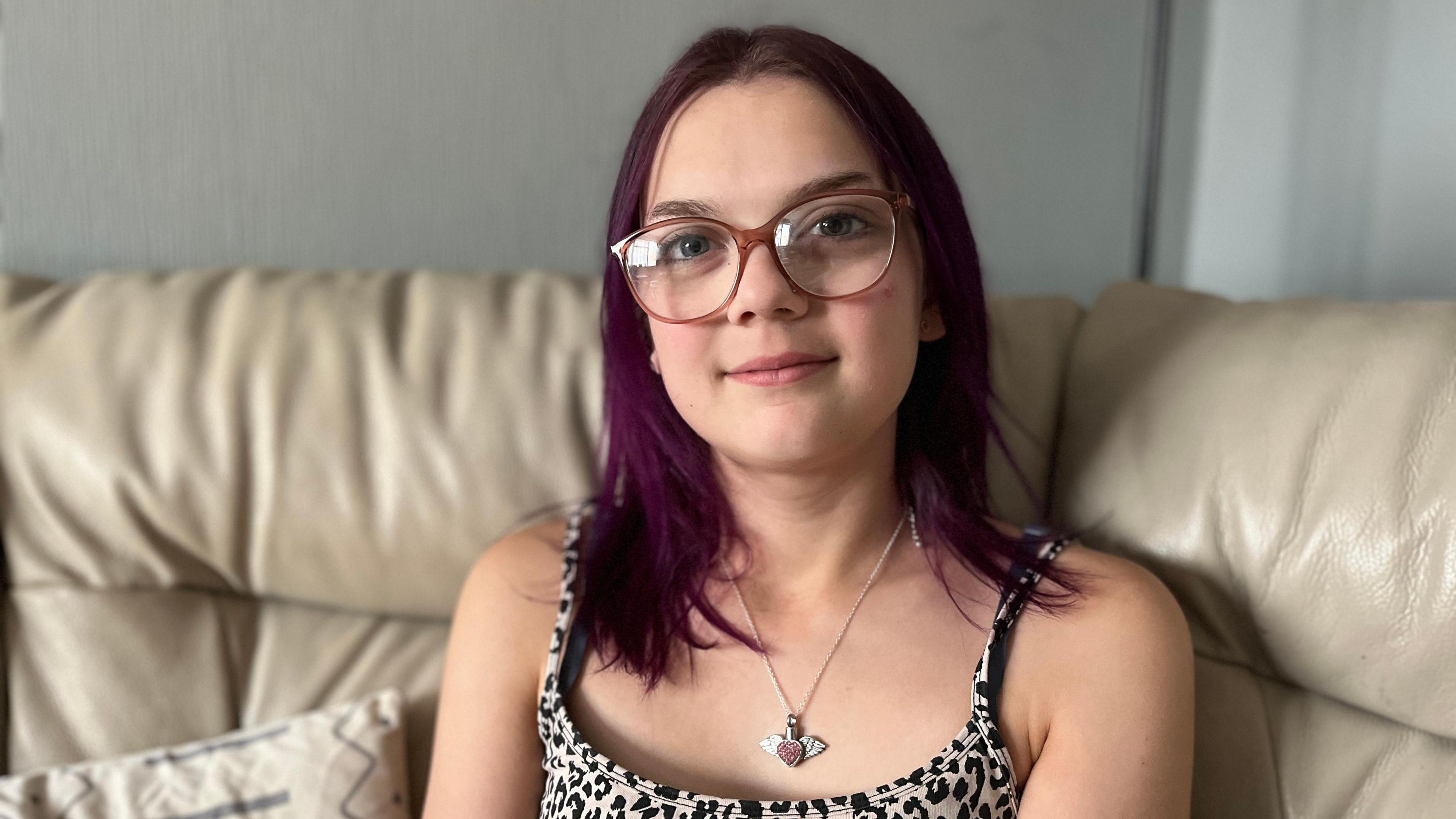 A teenage girl with glasses, purple hair and a heart necklace sitting on a sofa and smiling at the camera