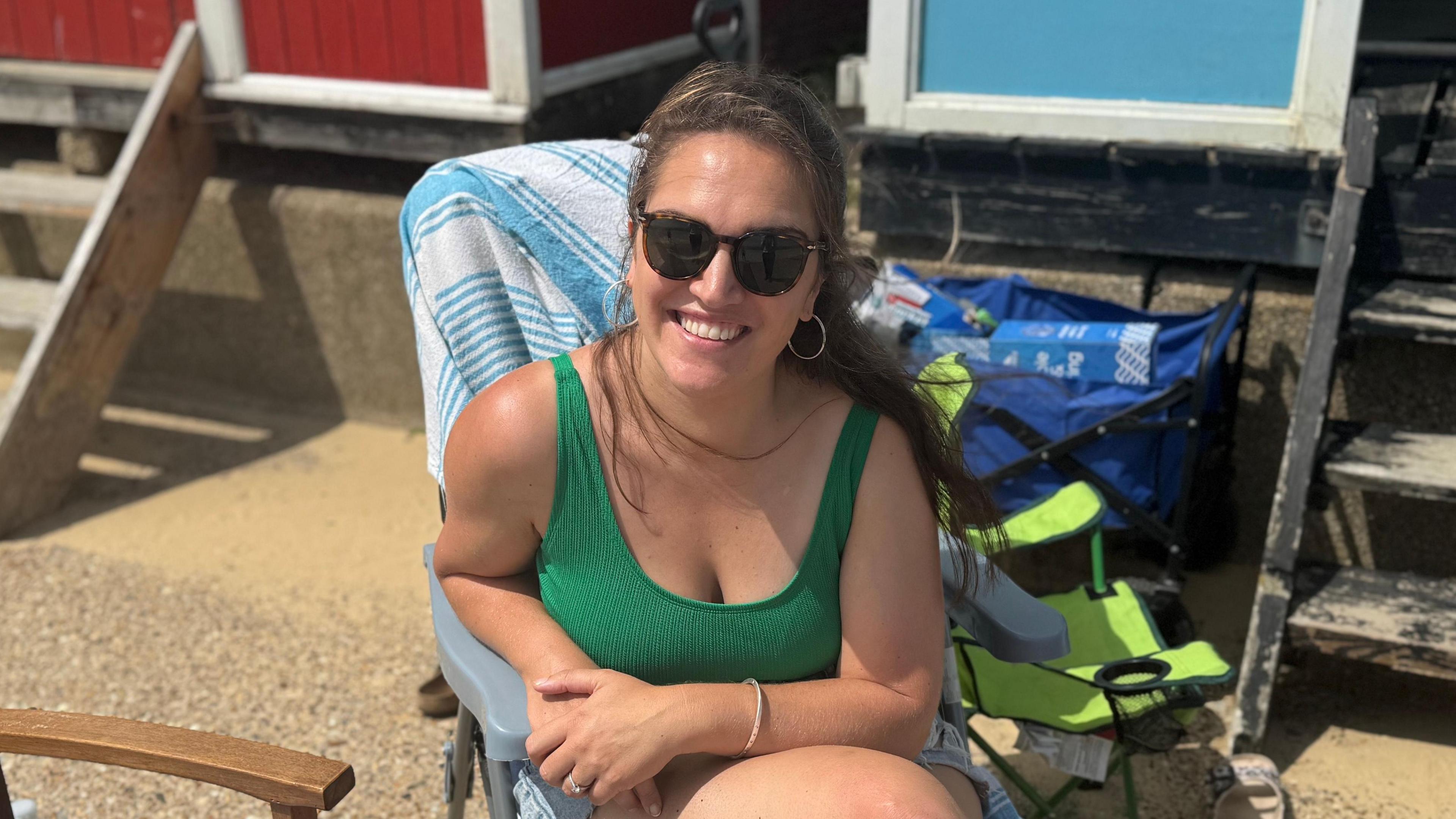 A head and shoulders image of Poppy Scale, who is wearing a green vest top and sitting in a beach chair outside a beach hut