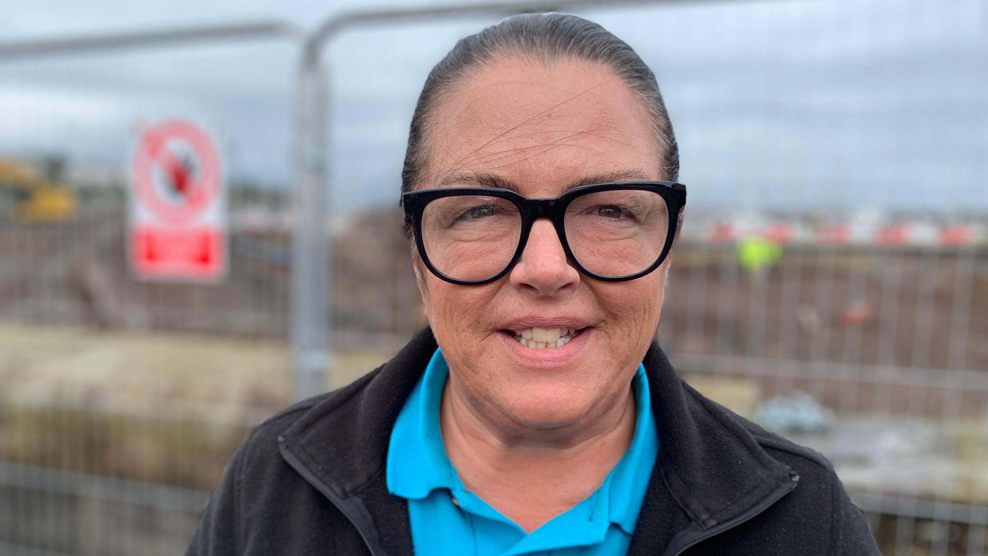 A woman with clear lensed black glasses smiles to the camera, wearing a black zip-up fleece with a blue polo shirt underneath. Behind her is a silver fence separating her and a building site, which is full of brown mud and several men in Hi-Viz jackets. 