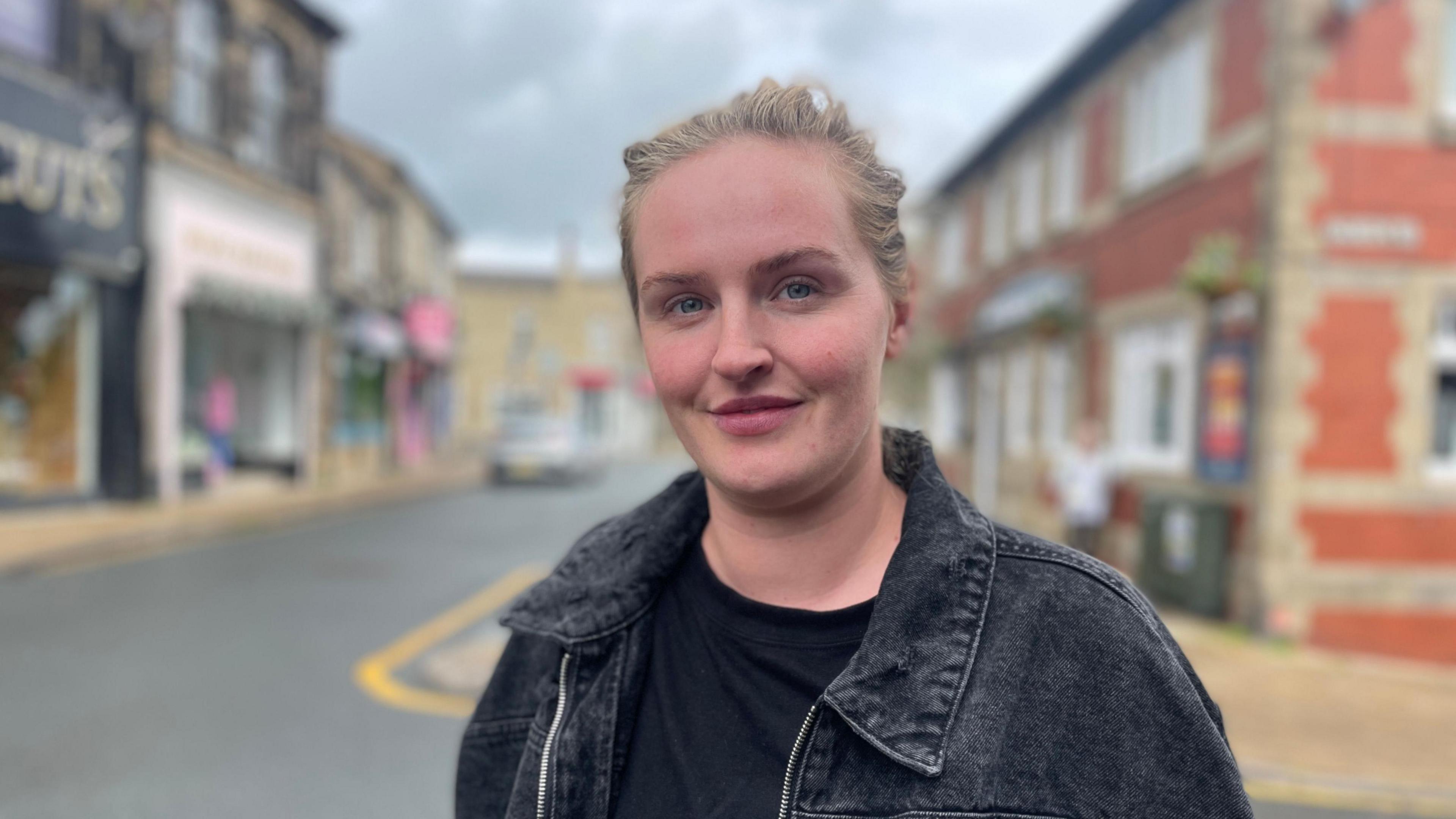 A lady is stood at a t- junction with her hair tied back wearing a black denim jacket with a black t shirt underneath.
Behind her is a row of shops on the left hand side and a red brick public house to her right.