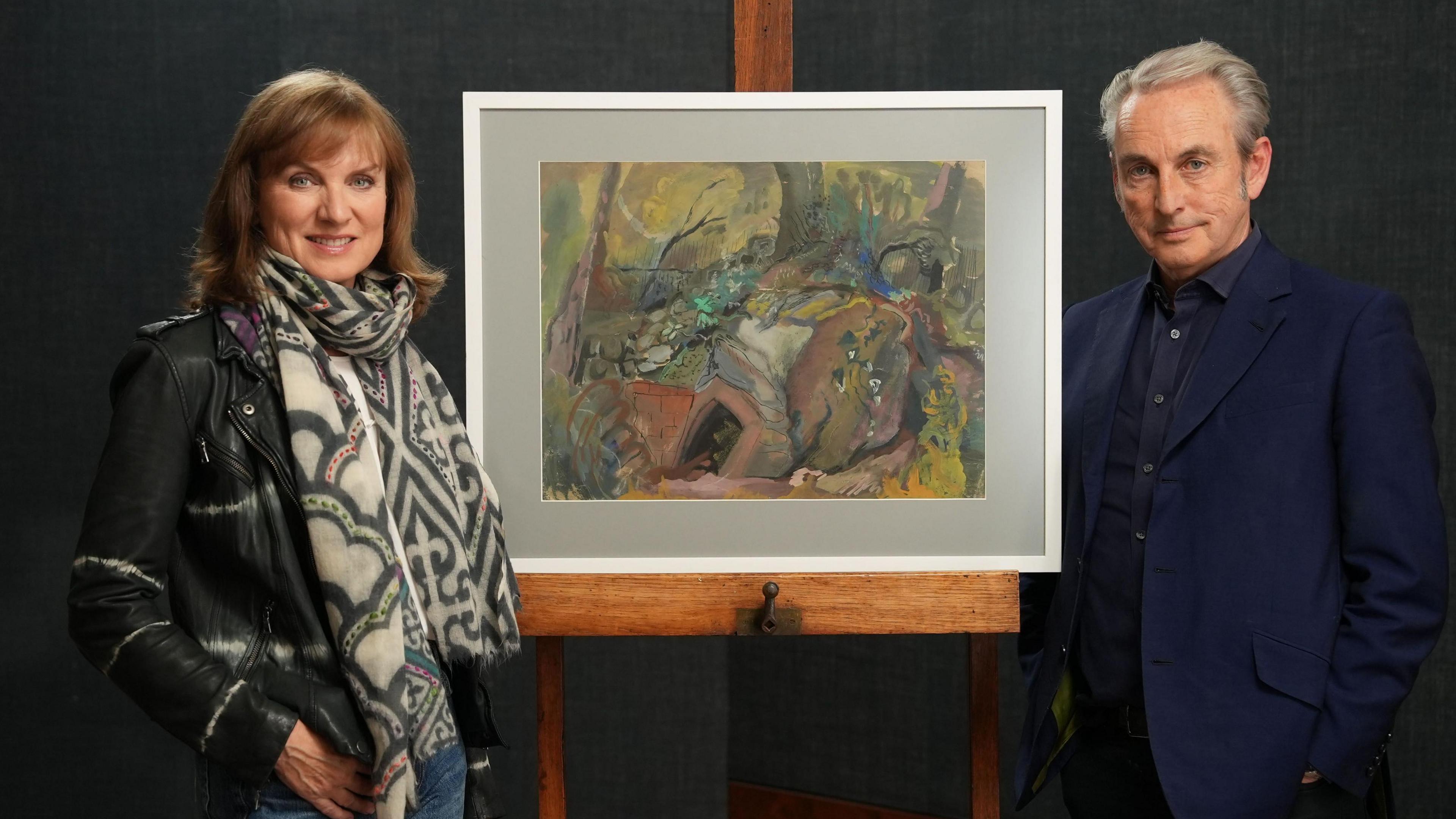 A man and a woman standing in front of a framed painting on an easel. She is wearing a black leather jacket and a patterned scarf. He is wearing a dark blue shirt and a blue suit jacket.