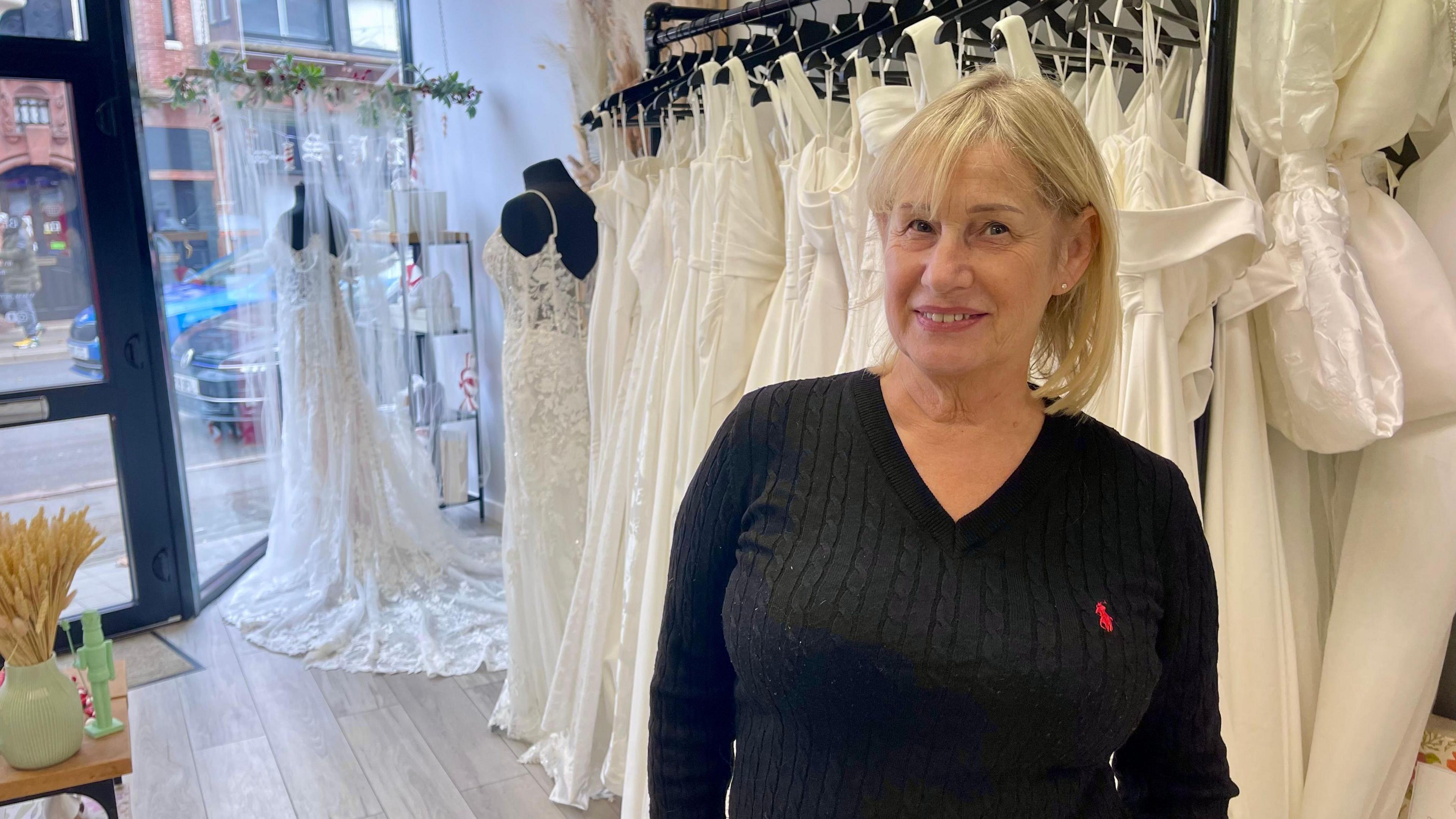 A woman in a black jumper and with short blonde hair is stood in a wedding dress shop. Behind her is a rack of around 30 white wedding dresses. There are two headless black mannequins at the front of the shop, each wearing a different white wedding dress. There is a also a stack of shelves, a small table with a green vase and a small flower display above the dress in the window. The street can be seen through the glass shopfront.