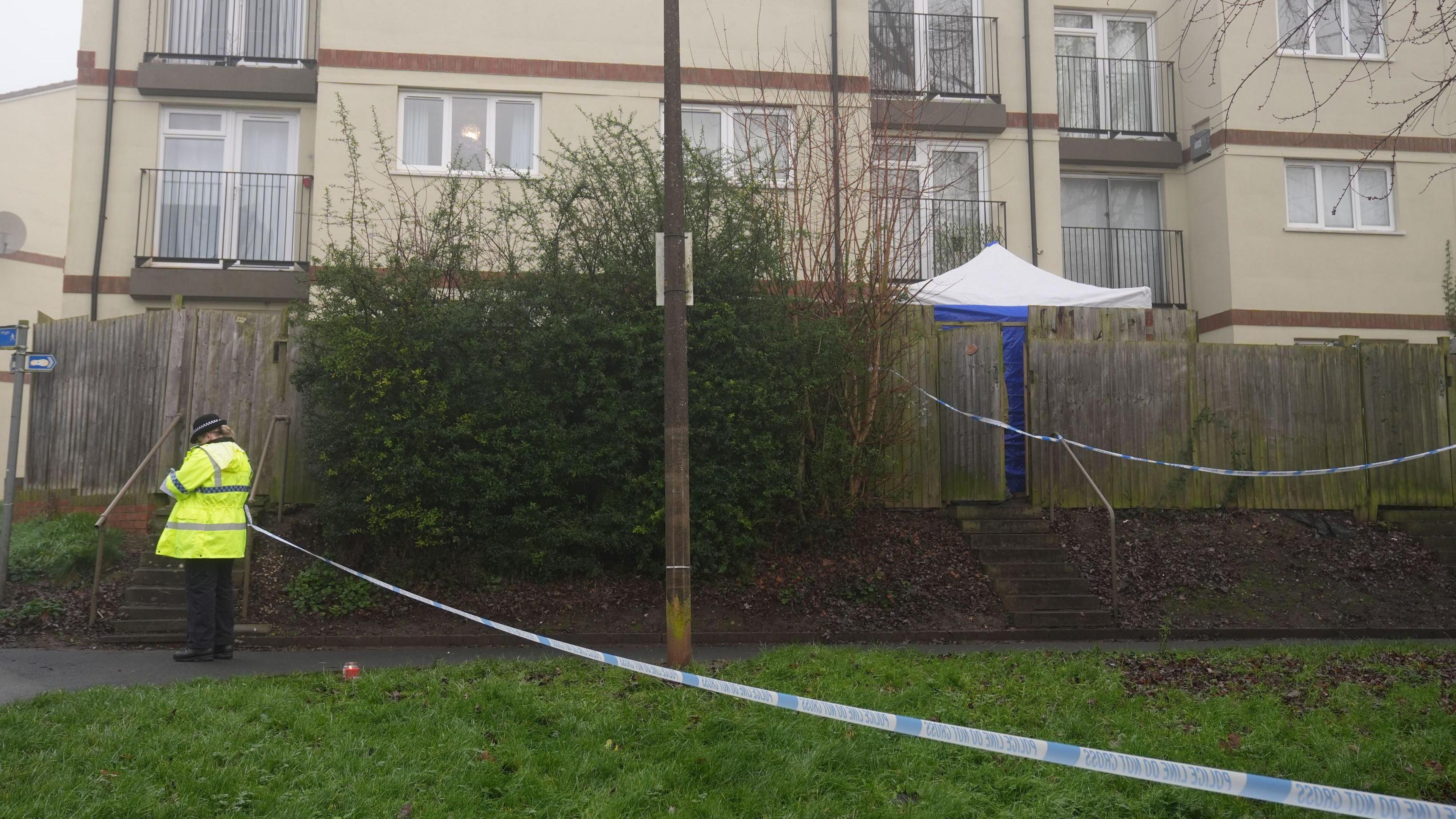 A police officer is stood by a police cordon. Within the taped-off area is a block of flats and a police forensics tent, positioned in an area behind wooden fence panels.