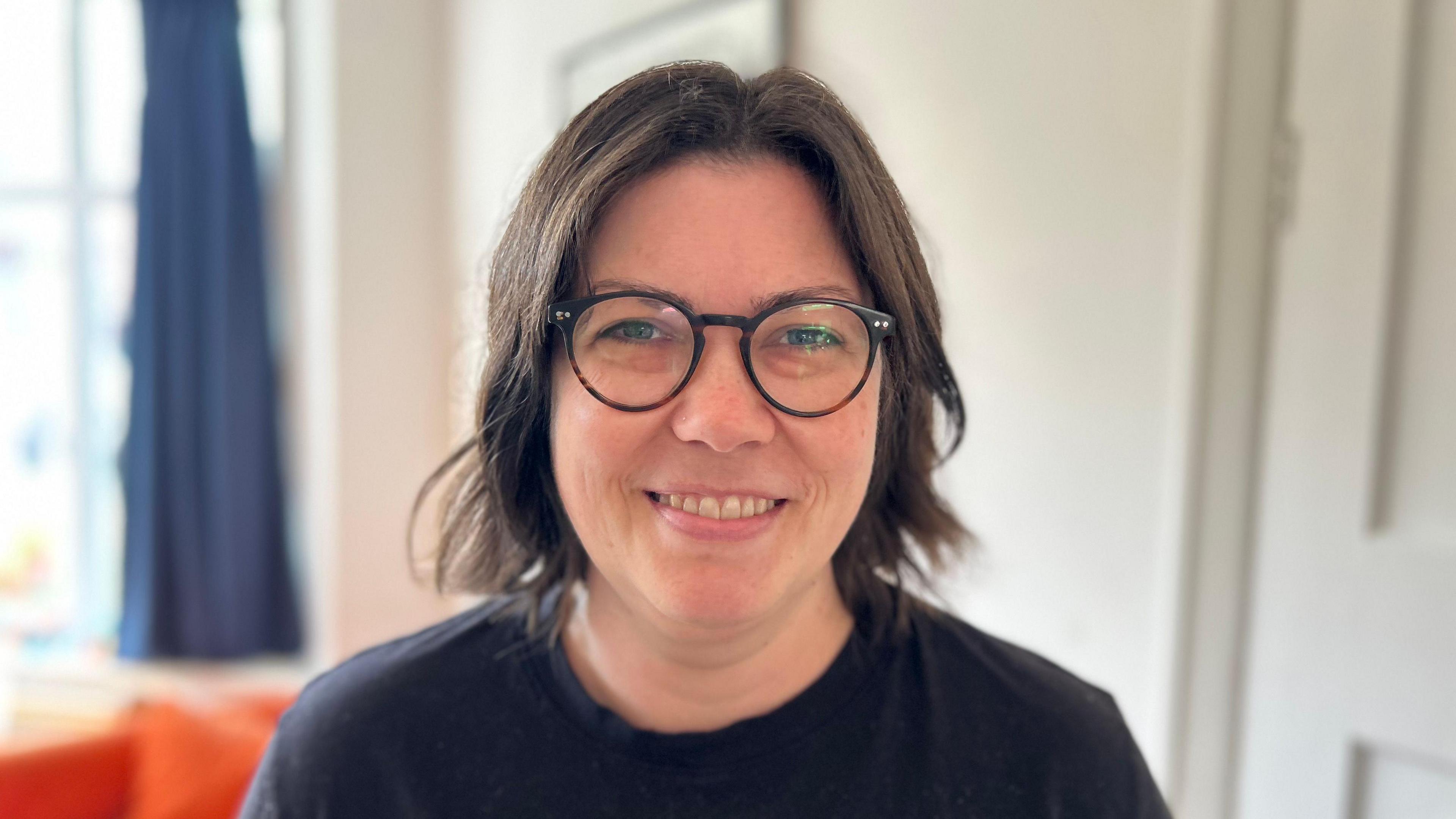 Molly Haworth, head and shoulders shot, showing a lady with dark hair and glasses smiling at the camera in a black t-shirt