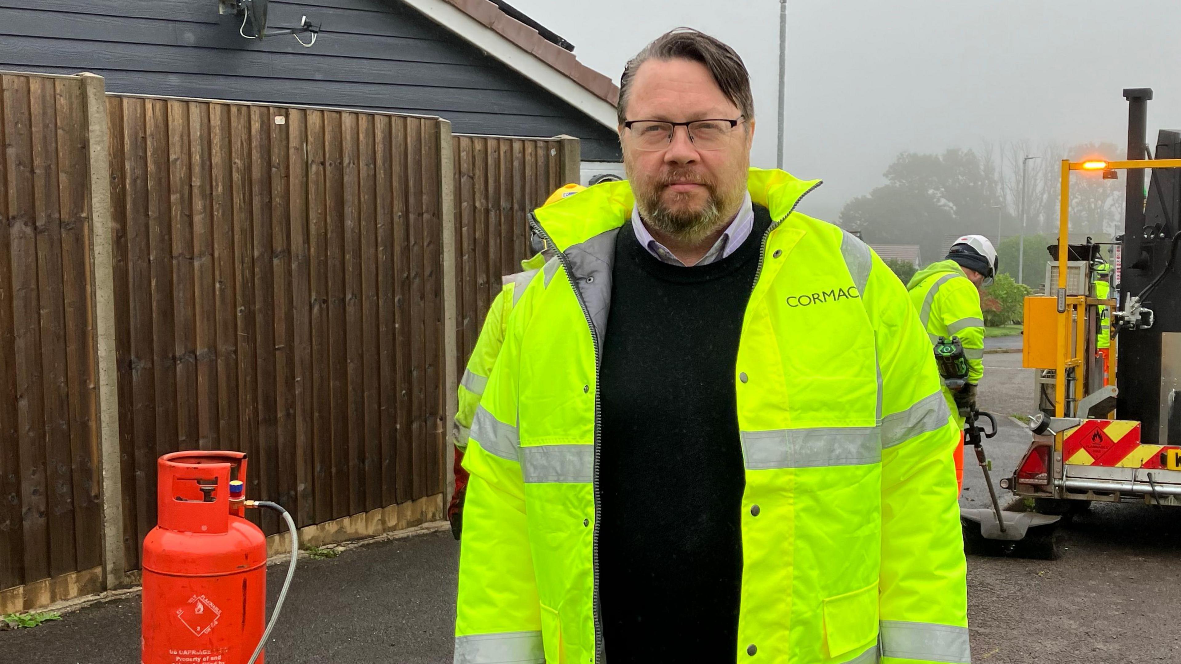 A man with in a shirt and black jumper wearing a high vis yellow jacket with a gas cannister to his right and workmen to his left standing in a residential area.